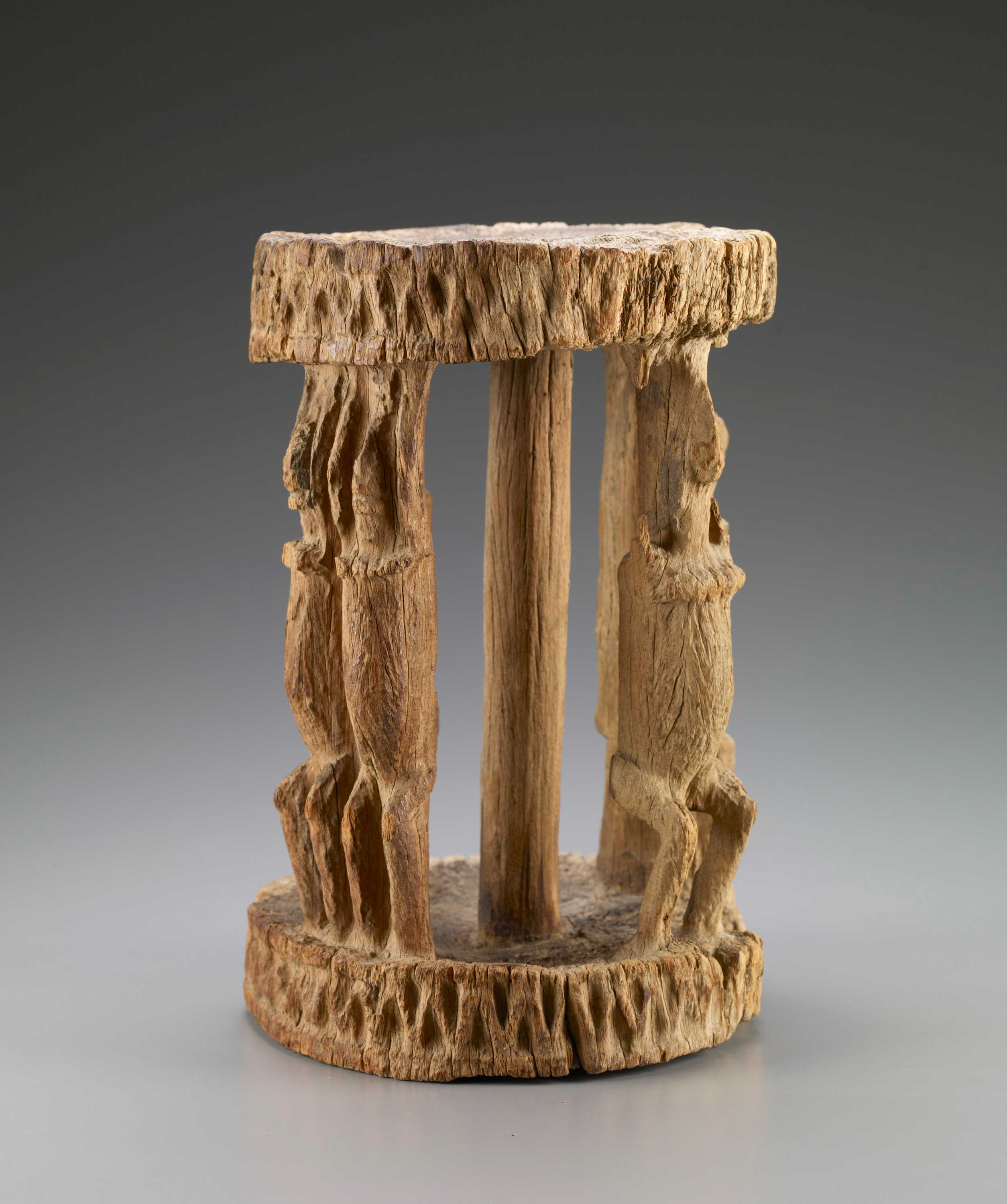 A wooden carved stool of three figures holding the top of the stool and carved patten into ends.