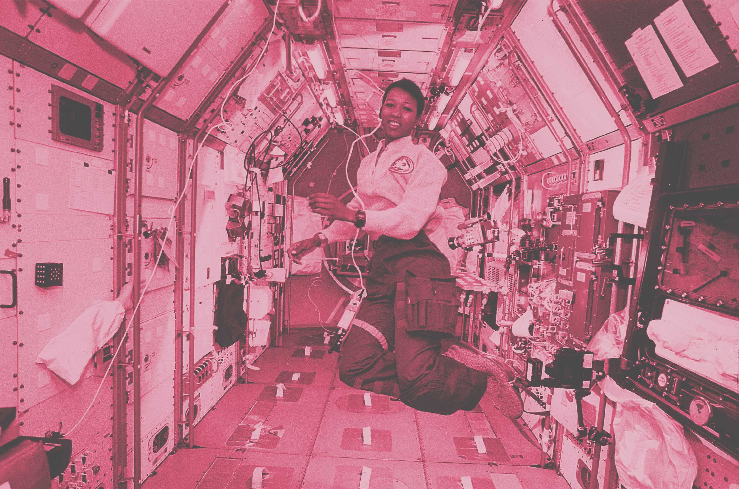 Astronaut Mae Jemison is onboard and floating inside a space craft. The image has a pink overlay.