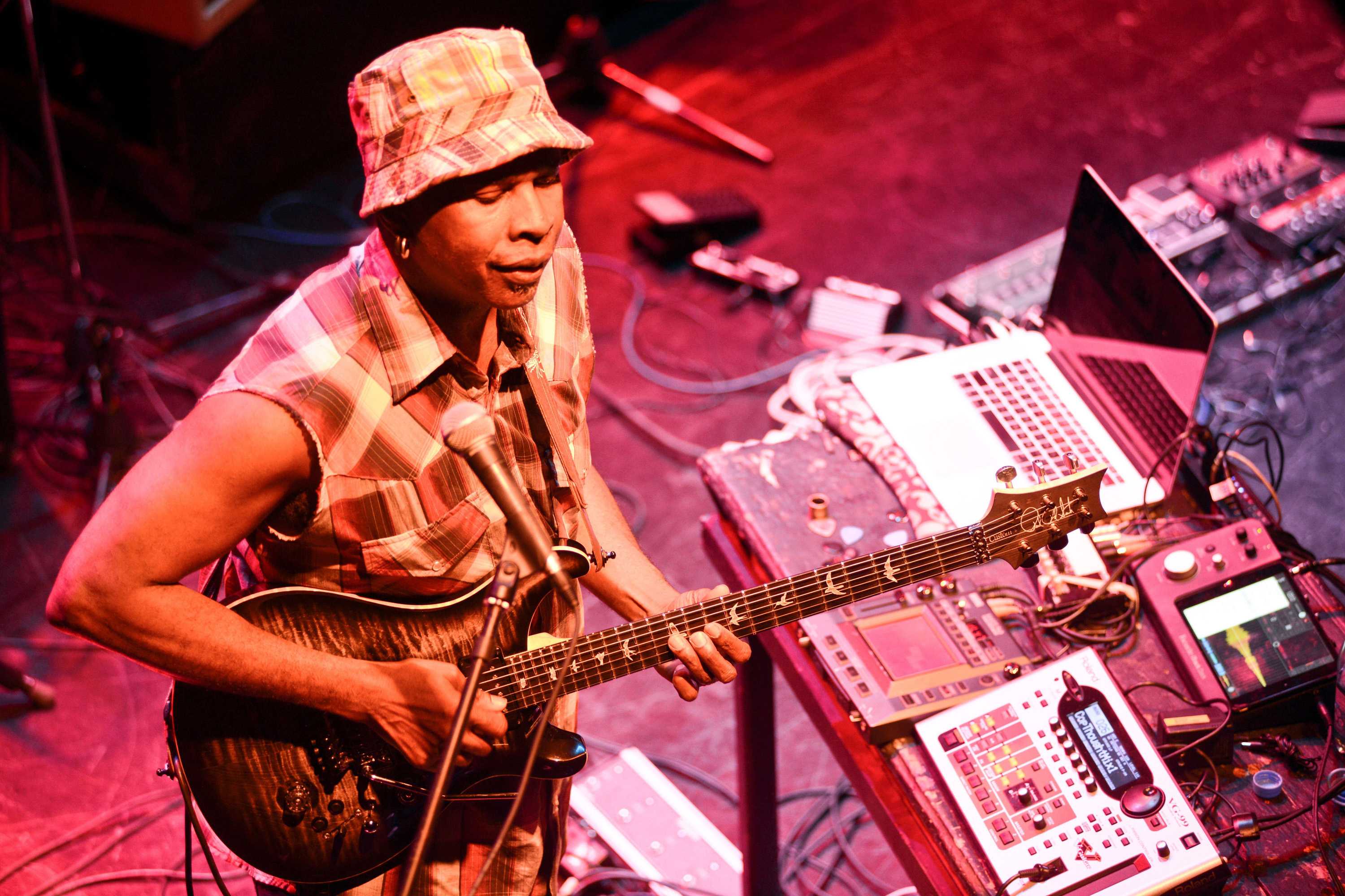 Vernon Reid singing into a microphone while playing the bass, surrounded by other equipment.