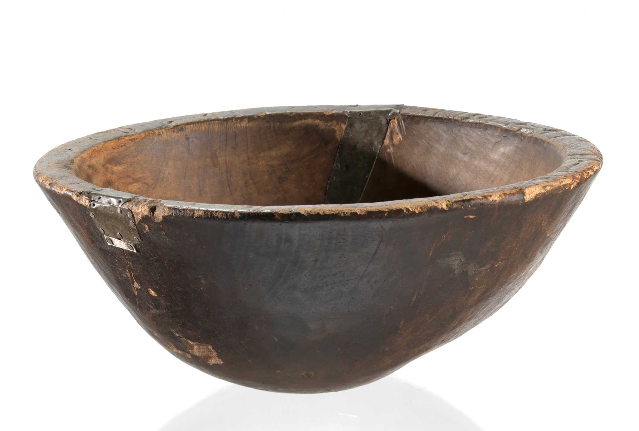 A large wooden bowl with an uneven shape and a rounded bottom. The lip is decorated with incised Adinkra symbols. Formed metal pieces were nailed over 2 separate cracks, acting as "patches." There is surface wear throughout, particularly on the bottom and around the lip.