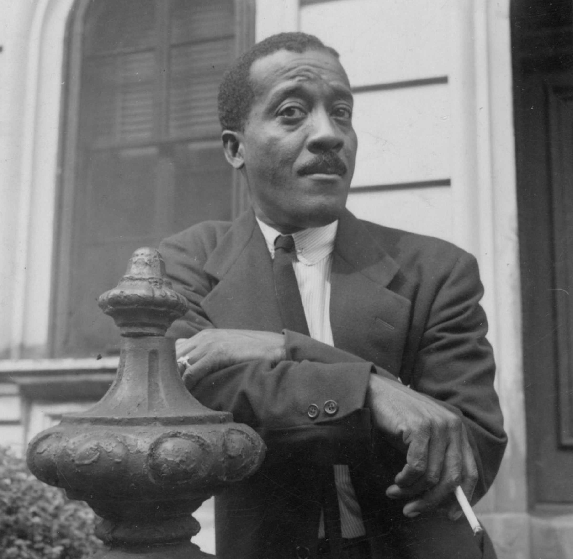 Black-and-white photo of a man in a suit leaning on a decorative metal post in front of a building.