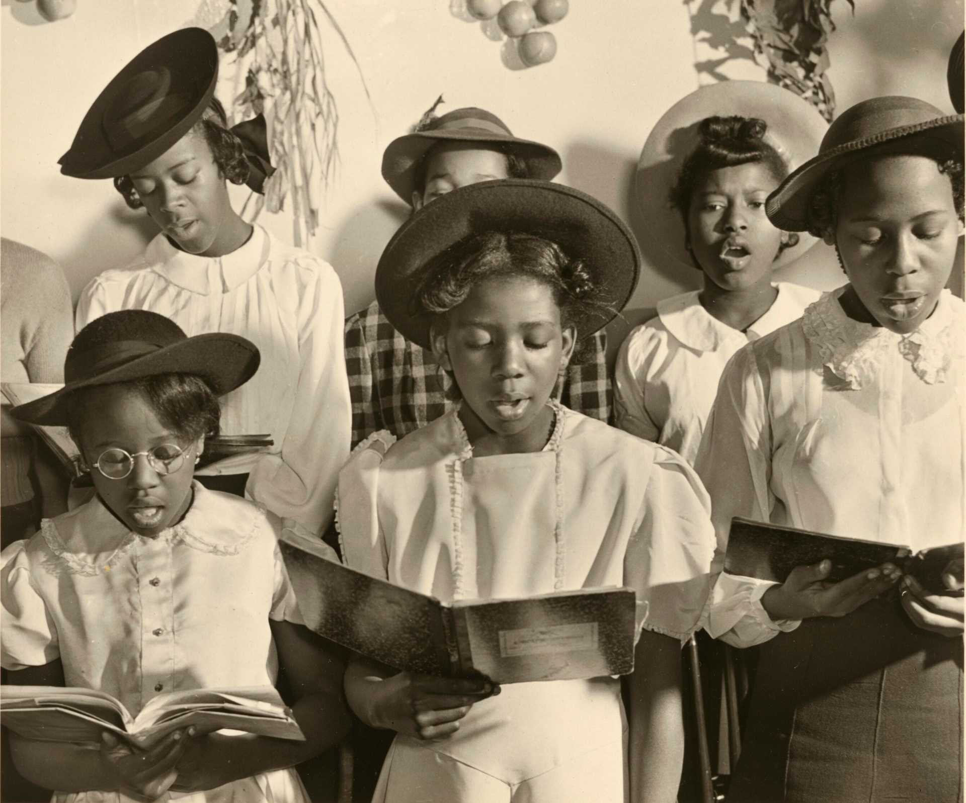 A black-and-white photograph of six girls wearing hats and singing from open books.