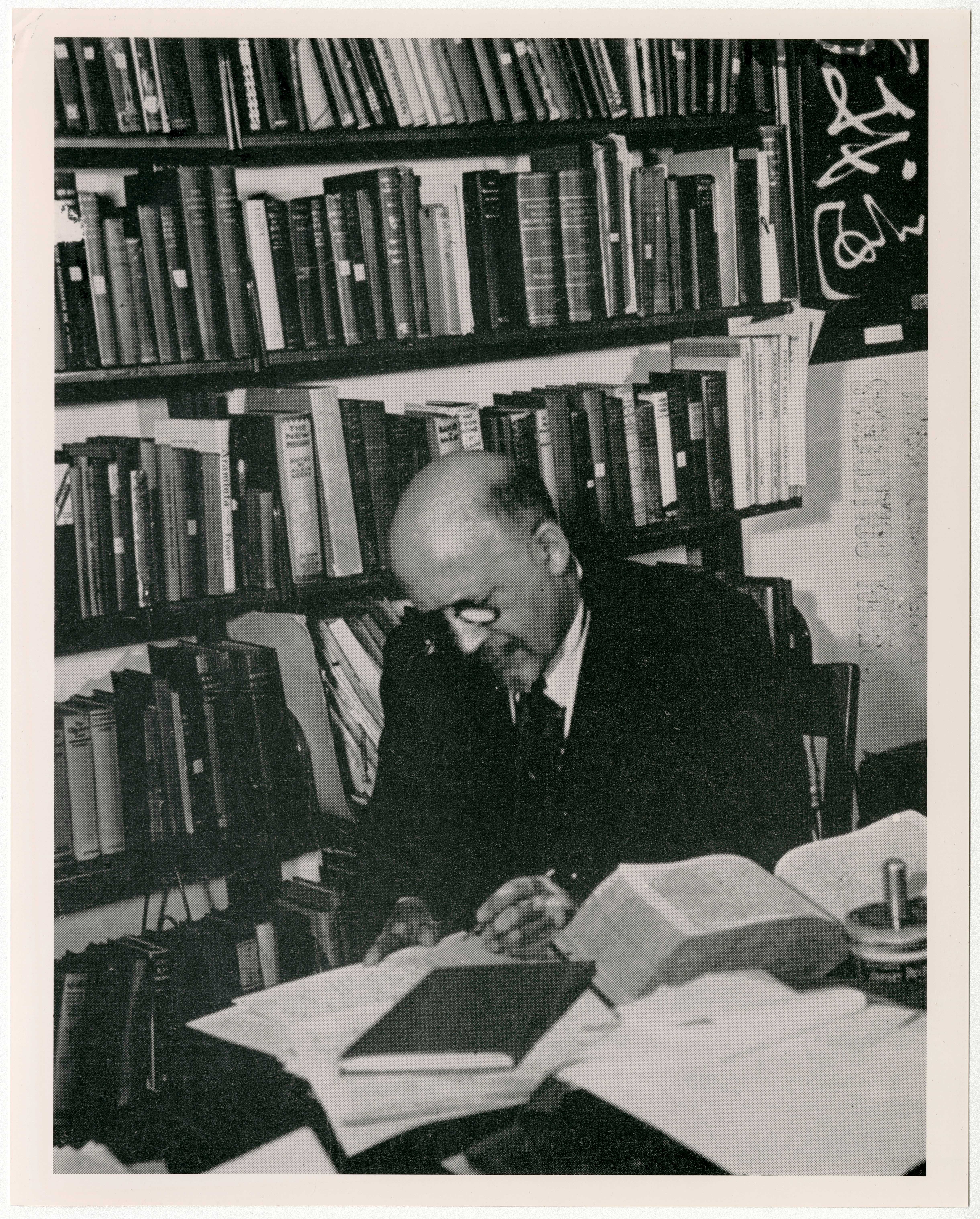 Black and white image of an W.E.B. Du Bois wearing glasses and a suit, seated at a desk cluttered with books and papers, surrounded by bookshelves in a study or office setting