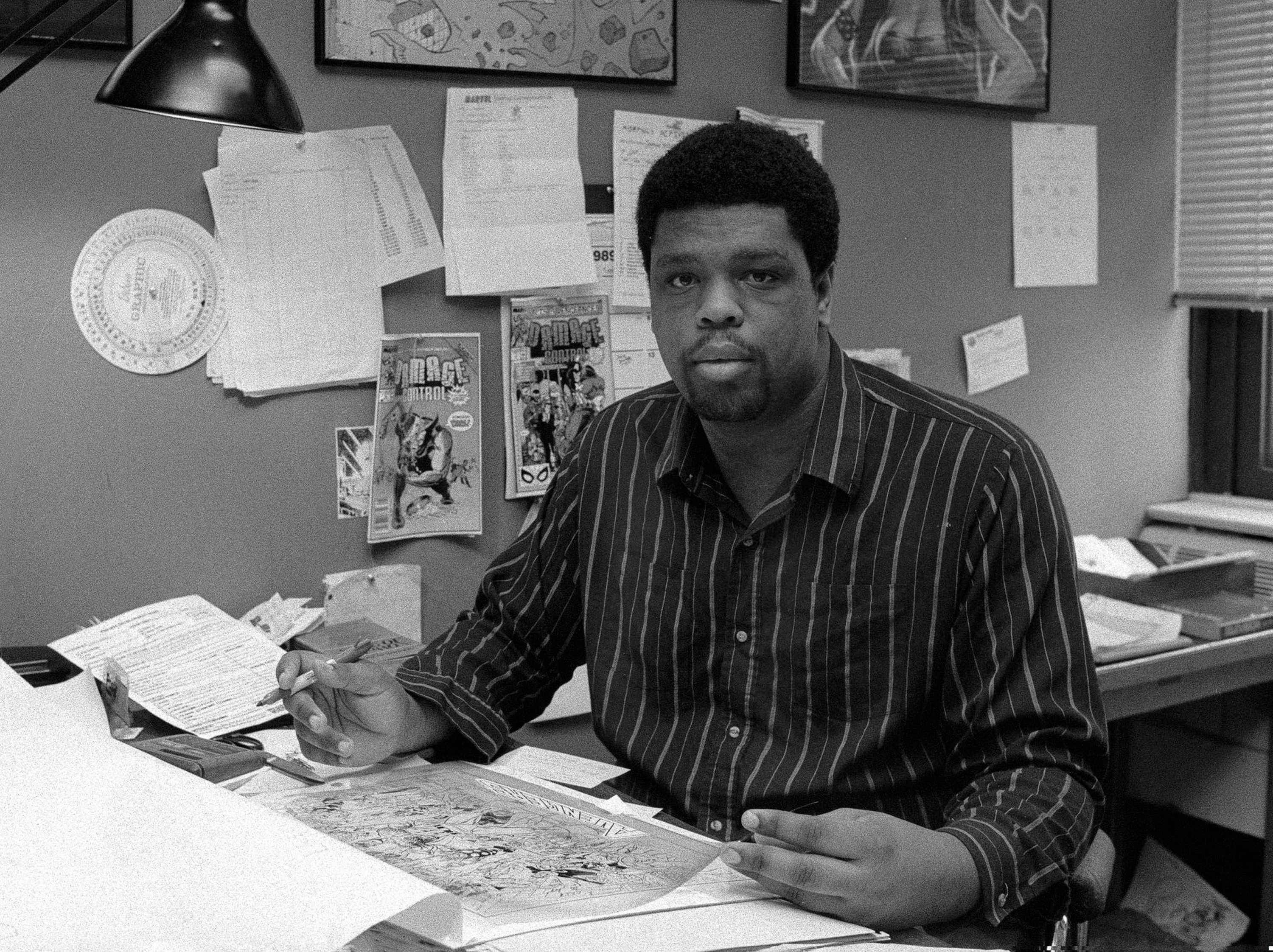 A black and white photo of Dwayne McDuffie working on a comic. He sitting at his desk in his office.