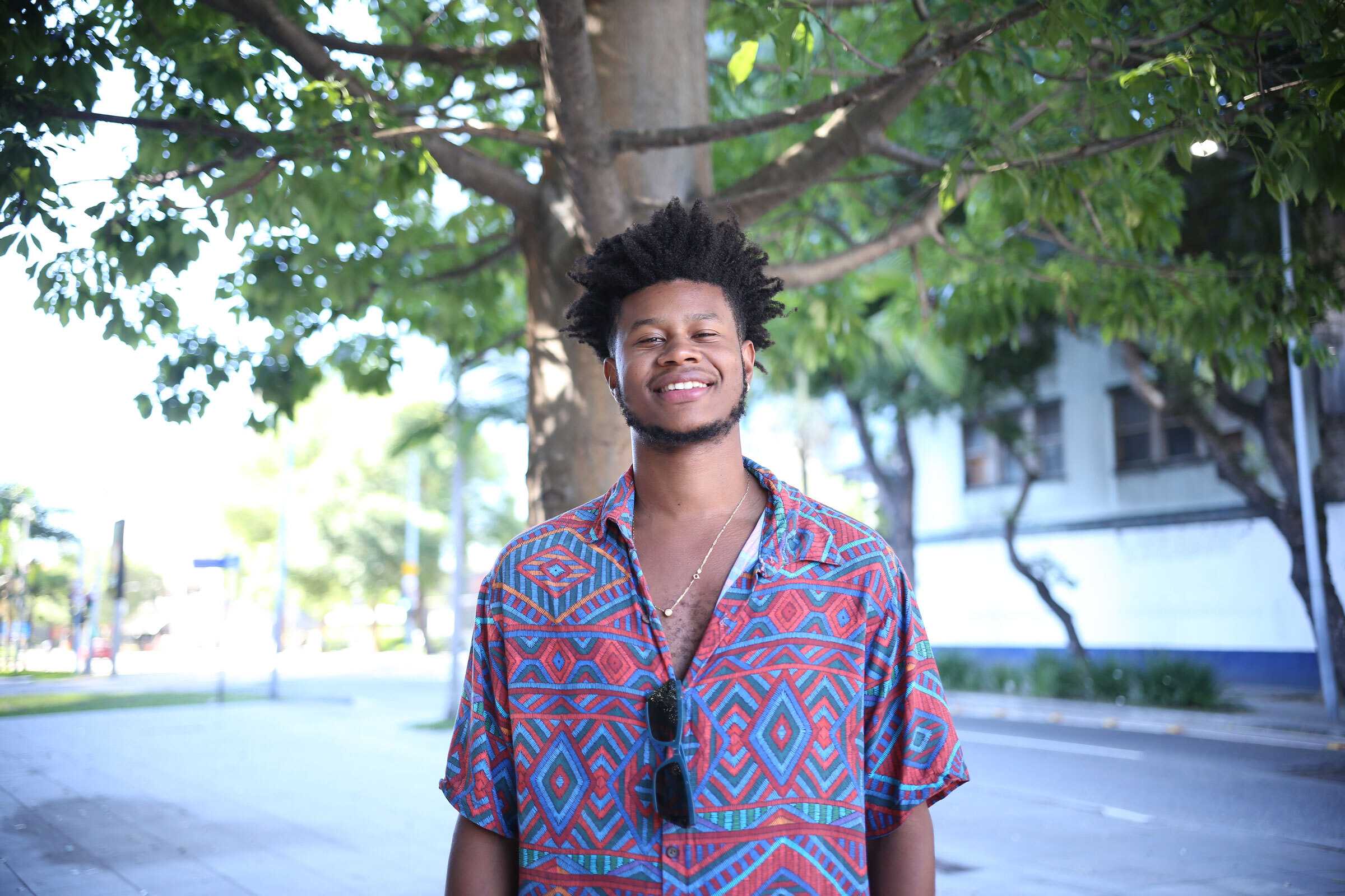 Person standing on a tree-lined city street, wearing a colorful geometric-patterned shirt with sunglasses hanging from the front.