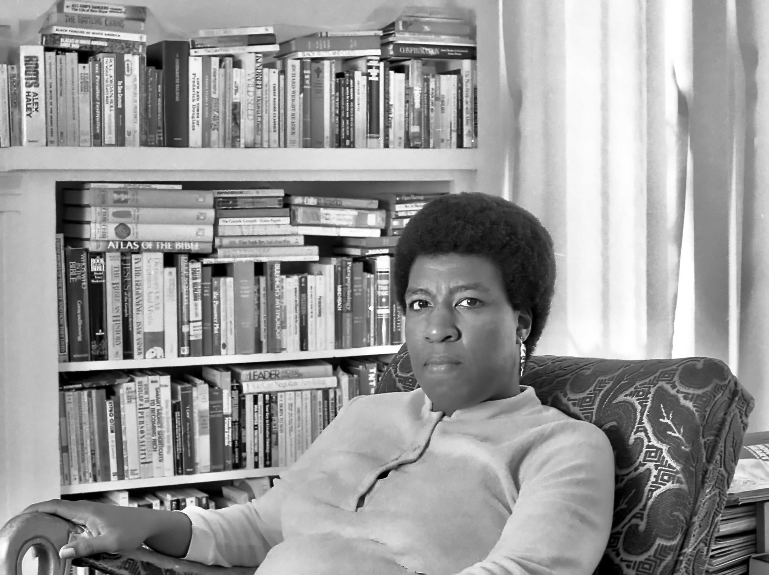 Black and white photograph of Octavia Butler sitting in front of her bookshelf.