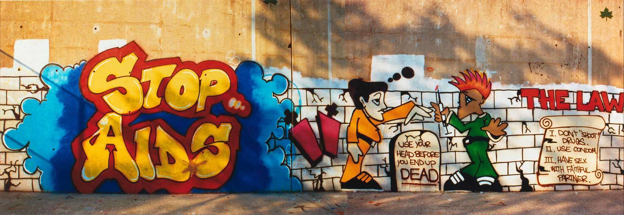 A color photograph of a portion of a Stop Aids graffiti mural in New York City. The mural is painted on a light tan wall. The bottom half of the image features a depiction of a white brick wall with cracks running through many of the bricks, running the full length of the image. A light and dark blue cloud features prominently on the left side of the image with the words [STOP / AIDS] spray painted in yellow letters outlined in red. The middle of the image features an illustration two men standing in profile, facing each other, on either side of a tombstone. The front of the tombstone has the text [USE YOUR / HEAD BEFORE / YOU END UP / DEAD.] written in black text. The word [DEAD] is underlined and the letters have drip lines giving it a bloody text effect. The man on the left is featured wearing a yellow shirt, yellow pants, and black shoes with yellow laces. He has three black thought bubbles above, and to the right of his head. He is holding his proper left hand splayed out, reaching for a syringe with a bloody hypodermic needle in the proper right hand of the man depicted on the right of the tombstone. The man on the right is depicted with red spikey hair, a green shirt, green pants and black shoes with white laces. On the far right of the image is the text [THE LAW] written in red block letters, outlined in black, above a depiction of scrolled paper with the text [I. DON’T “SHOOT” / DRUGS. / II. USE CONDOM. / III. HAVE SEX WITH FAiTHFUL / PARTNER.] written in black text. There are no inscriptions on the recto. On the verso the image is signed in blue ink by the photographer.