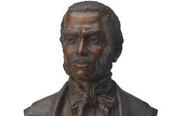 This bronze portrait bust depicts Frank McWorter, formerly enslaved American and founder of New Philadelphia, Illinois. Shown from the chest up, the bust is mounted on a black stone rectangular base with the front protruding slightly at an angle. Frank McWorter wears a coat with a swirling texture incised onto the surface. under the coat is a buttoned waistcoat, shirt, high collar and cravat. He has a mustache and sideburns, and his head is turned slightly to the viewer’s' left. The bust is signed by the artist on the back of the left shoulder.