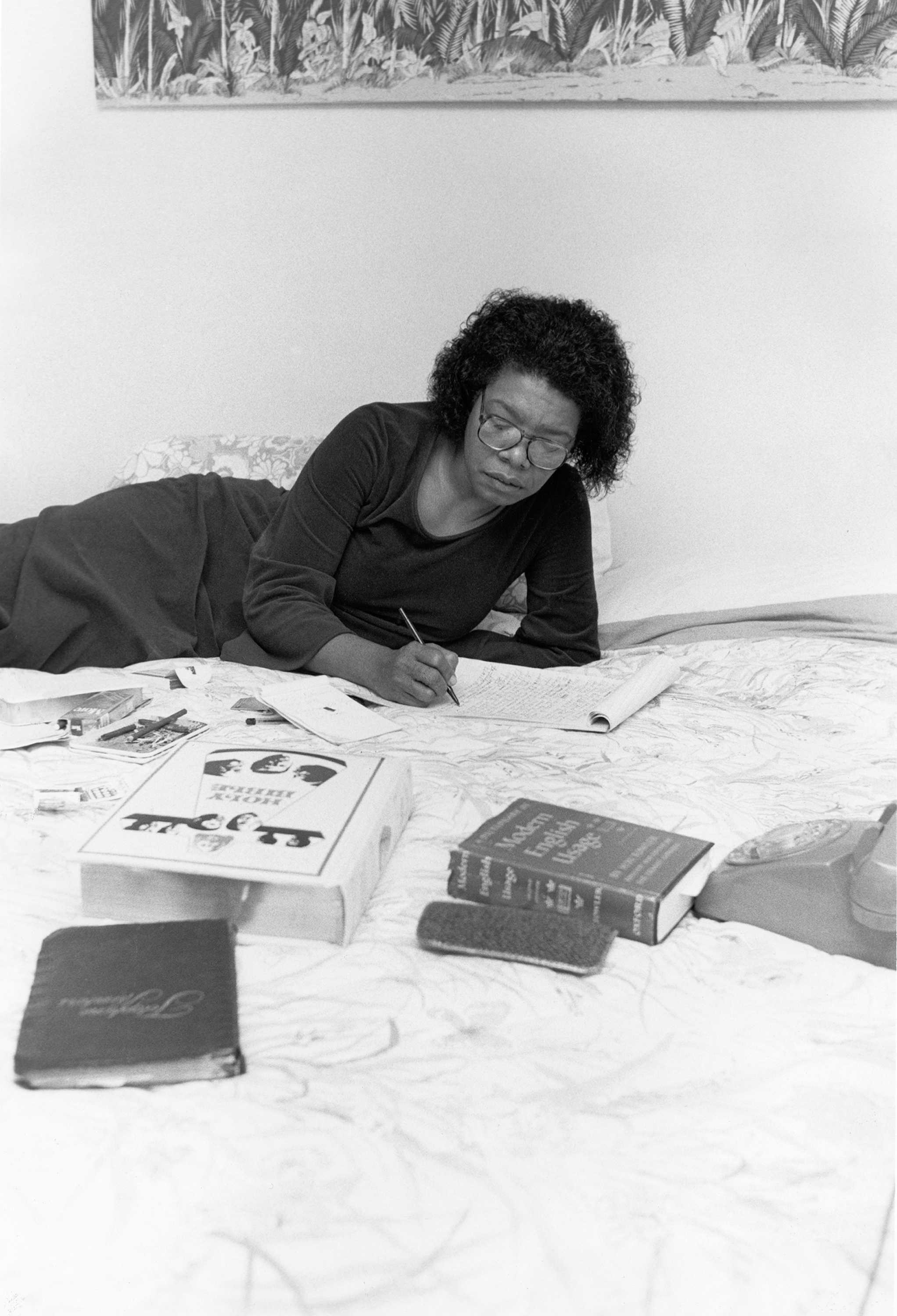 Black and white photograph of Maya Angelou working on her bed with a thesaurus, a dictionary, and the Holy Bible