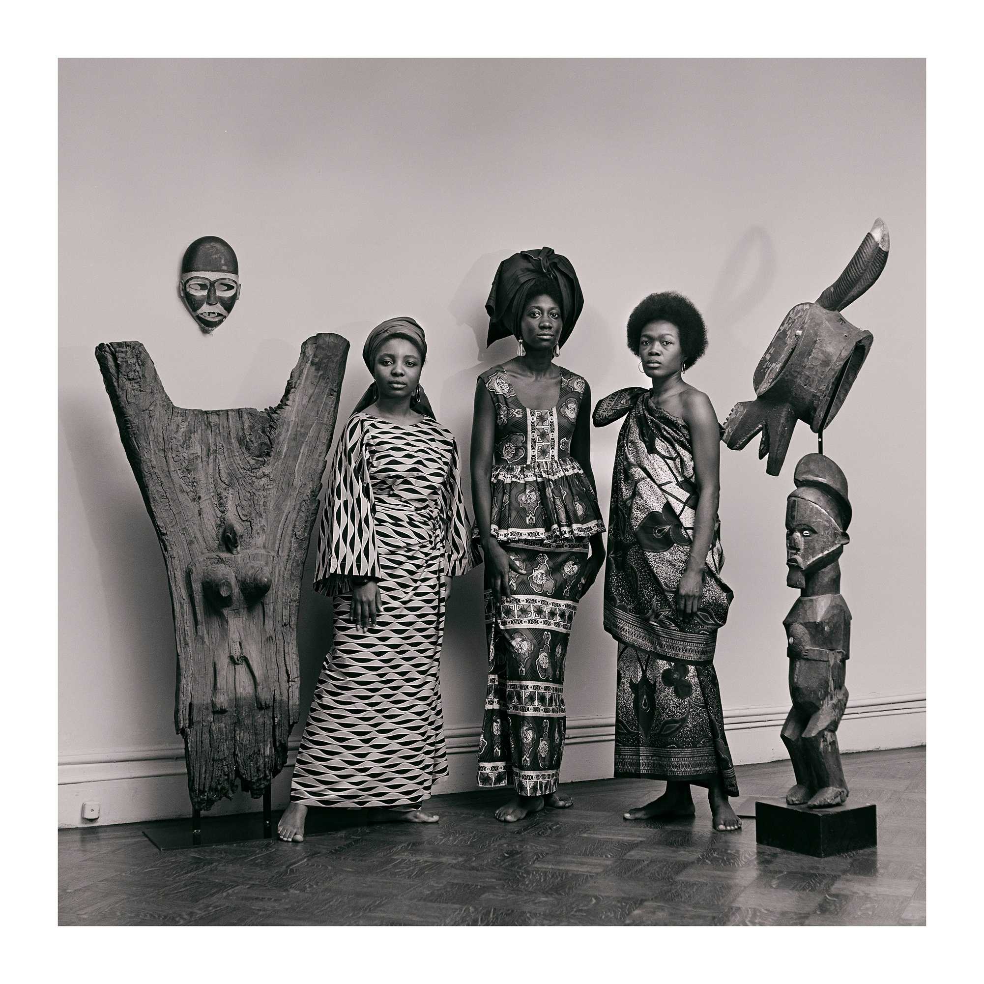 In a black and white photograph, models stand in between two wood statues.