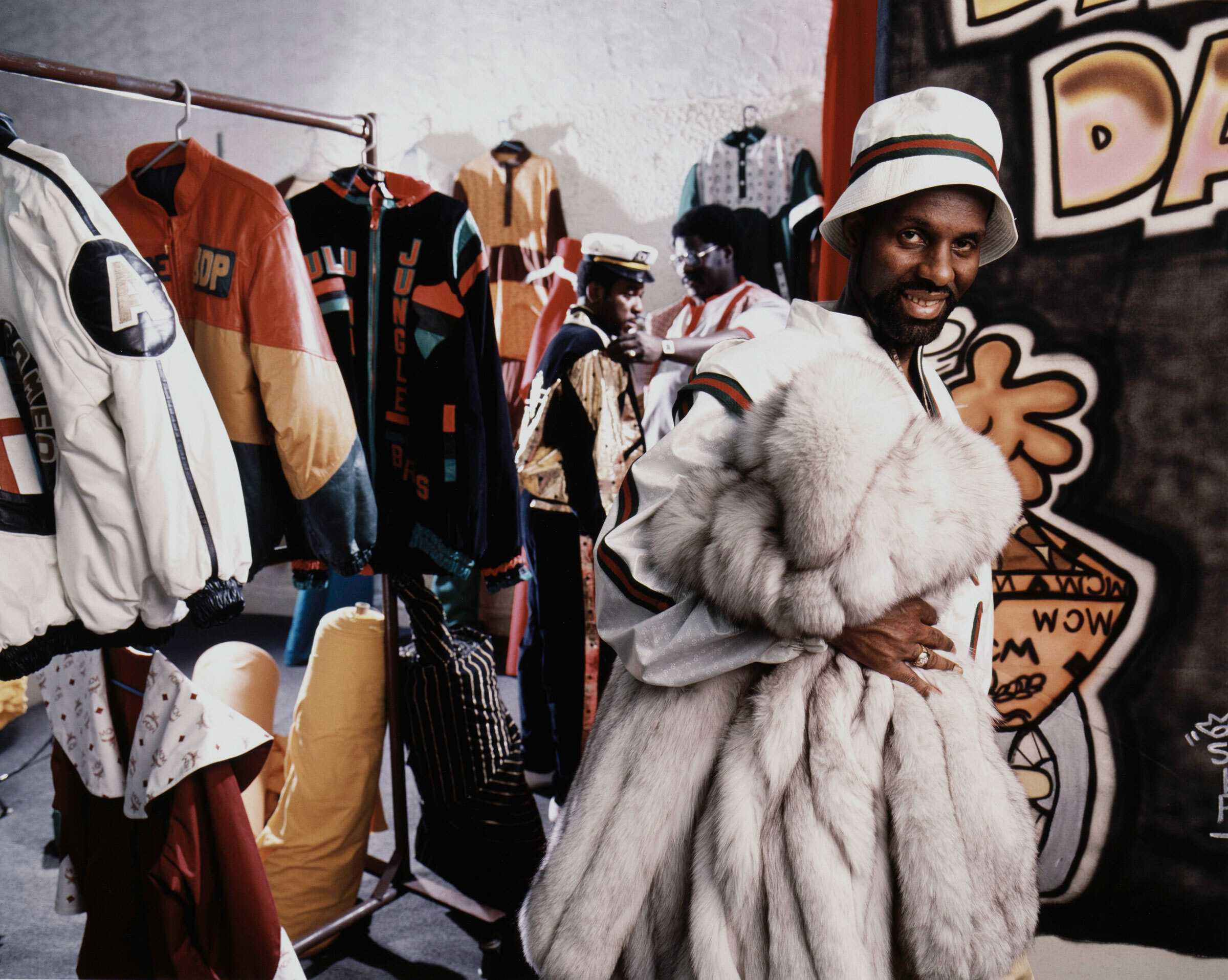 Color photograph depicting Dapper Dan in his clothing boutique in Harlem, New York. Dan is pictured just right of center, clutching a white fur coat and looking directly at the camera.
