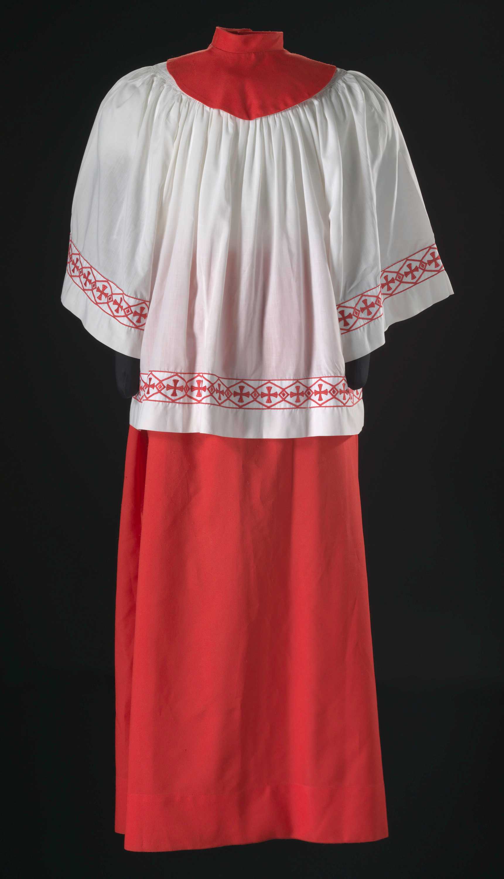 Short, white and red acolyte robe (a), with skirt (b), and collar (c). The robe has a border along the waist and sleeves. Border is a red embroidered geometric pattern with interlocking, elongated diamonds. Within each diamond is a cross. Where the diamonds overlap is a smaller solid diamond. Robe length stops just below the waist. Long, red, adjustable skirt with ties at back. Hem runs roughly four inches from the bottom. String ties for adjustable waist. Red collar with round base stitched to the collar band. Collar is secured with two (2) snaps at the back below the collar band.