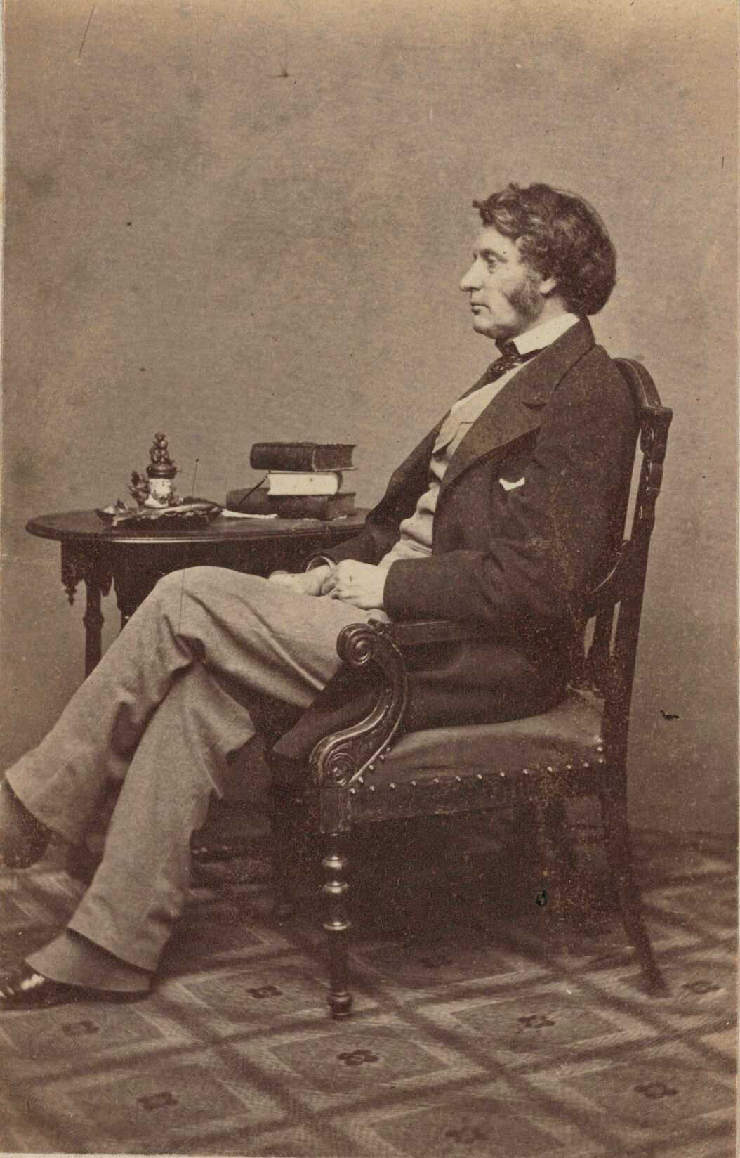 Carte-de-visite of Charles Sumner in full length seated profile. Sumner is pictured with his left profile facing the camera. His hands are resting in his lap and his left leg is crossed over his right leg at the knee. He is wearing a light colored vest and trousers with a white shirt, dark tie, and dark jacket. Spats peak out below his pant legs, partially covering his dark colored low-heeled shoes. His hair is moderately long and he has long sideburns. Sumner is seated in a carved armchair with an upholstered seat with a round wooden side table behind him. A stack of books, some loose papers, and possibly an inkstand are placed on the table. The floor of the room is covered in a geometric patterned carpet. There is a double-lined border printed in gold ink surrounding the outside edges of the card mount.

Printed on the back of the photograph is  E. and H.T. Anthony's mark.

The photograph is housed in the album 2017.30. The album page has a triple-lined, gold border framing the print. Handwritten in graphite in the bottom of the printed frame of the window on the album page is the text "Charles Sumner". Handwritten in the lower left corner of the album page in graphite is the name "E ANTHONY-BRADY".
