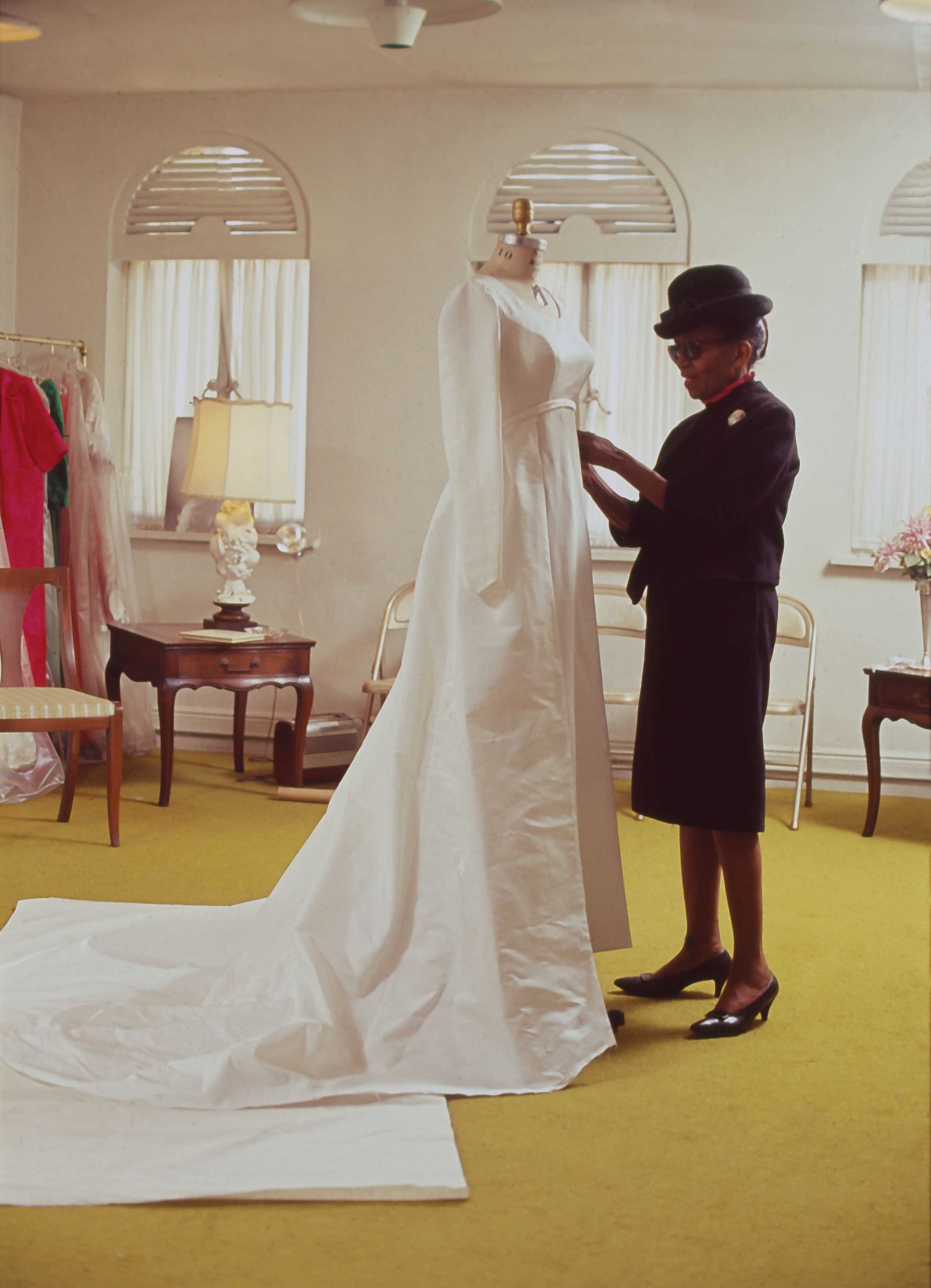 A photograph of Ann Lowe is pinning a wedding gown while wearing an classic all black dress suit.