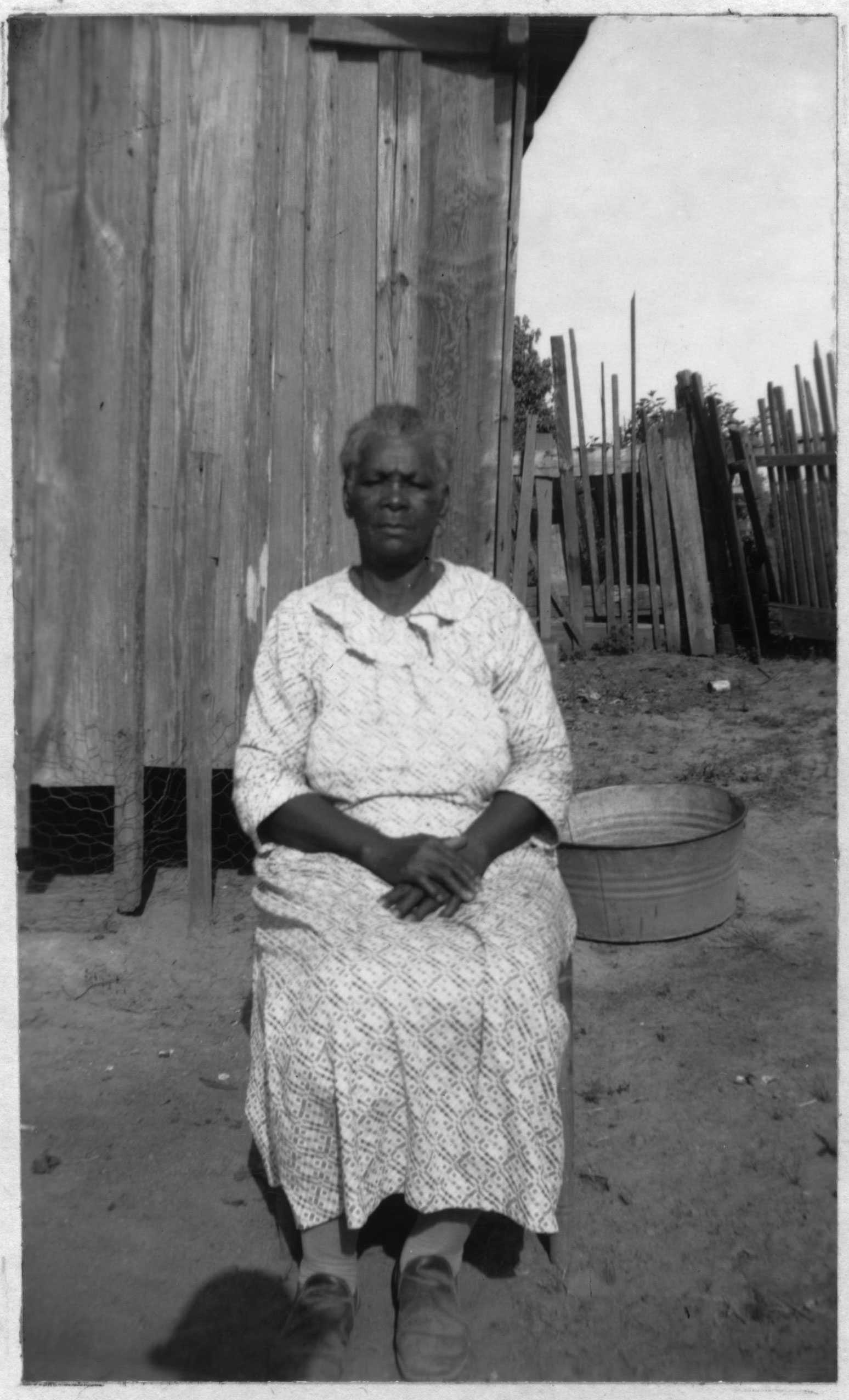 Elderly woman sitting in front of a wooden building, wearing a patterned dress, with a washtub beside her.