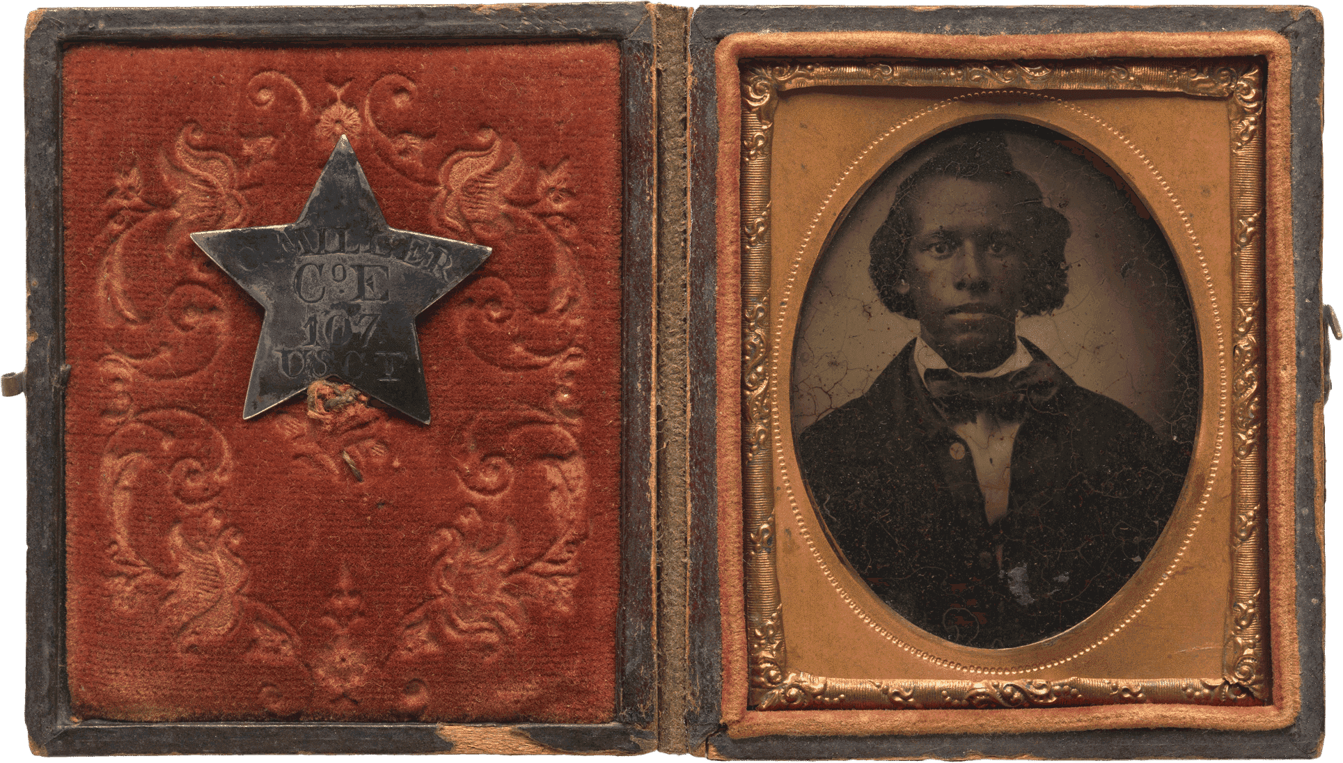 Tintype of Creed Miller, a soldier of the Kentucky 107th Regiment, Company E (and later Company C), United States Colored Troops. The tintype is encased in a copper scrolled frame with an oval window within a red velvet-lined, brown leather box. The tintype depicts Creed from the waist up in a dark overcoat, white collared shirt and bow-tie. His hair is styled and parted to the side. His miltary identification pin is fastened to the red velvet fabric on the interior of the case. The pin is a silver five pointed star with  [C. MILLER / Co. E / 107 / USCT] engraved at center. The front and back of the case is brown leather with detailed scroll work consisting of vines and flowers. The case has a metal hook and eye closure.