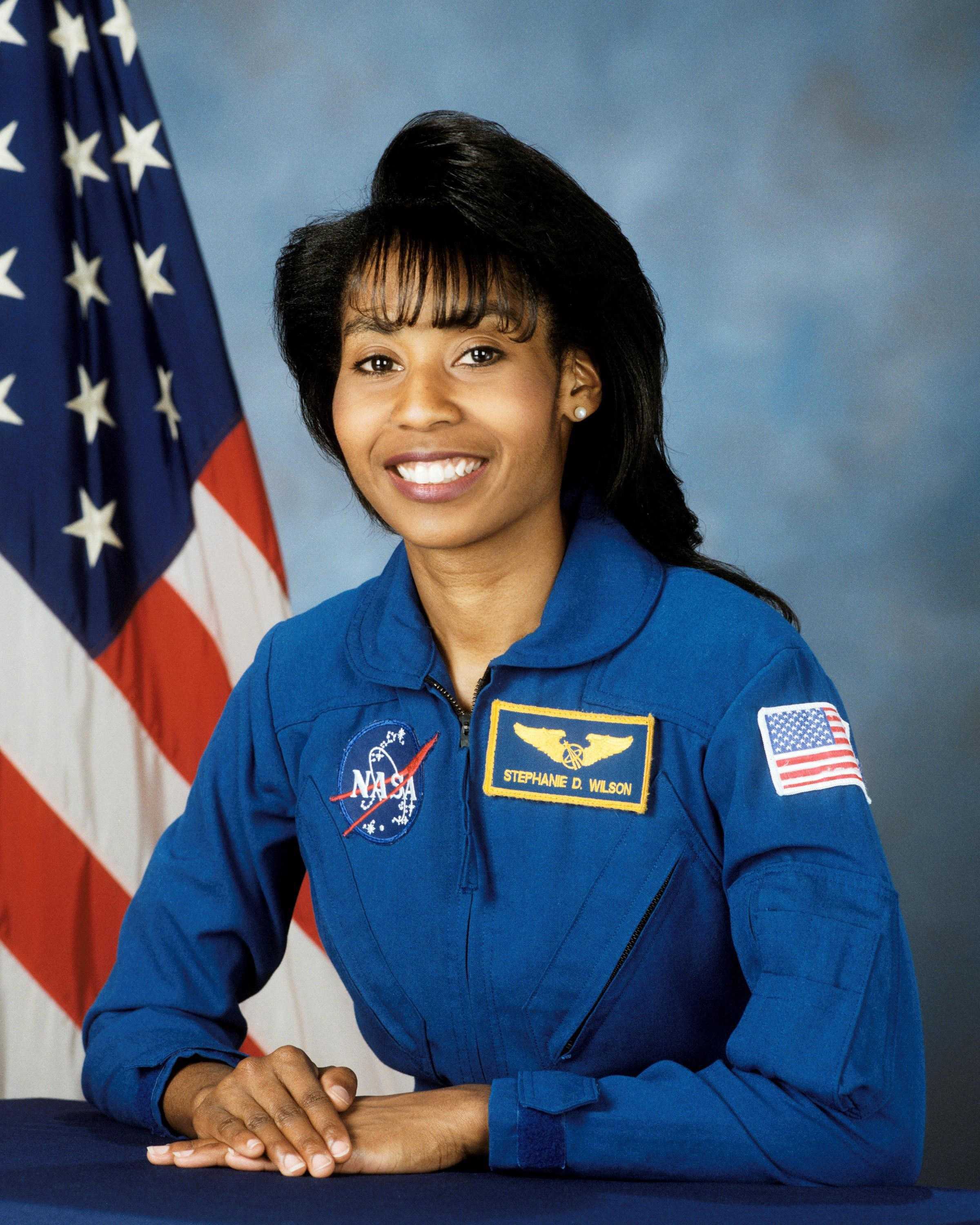 Wilson is wearing a blue NASA uniform and is sitting with her hands crossed on a table.