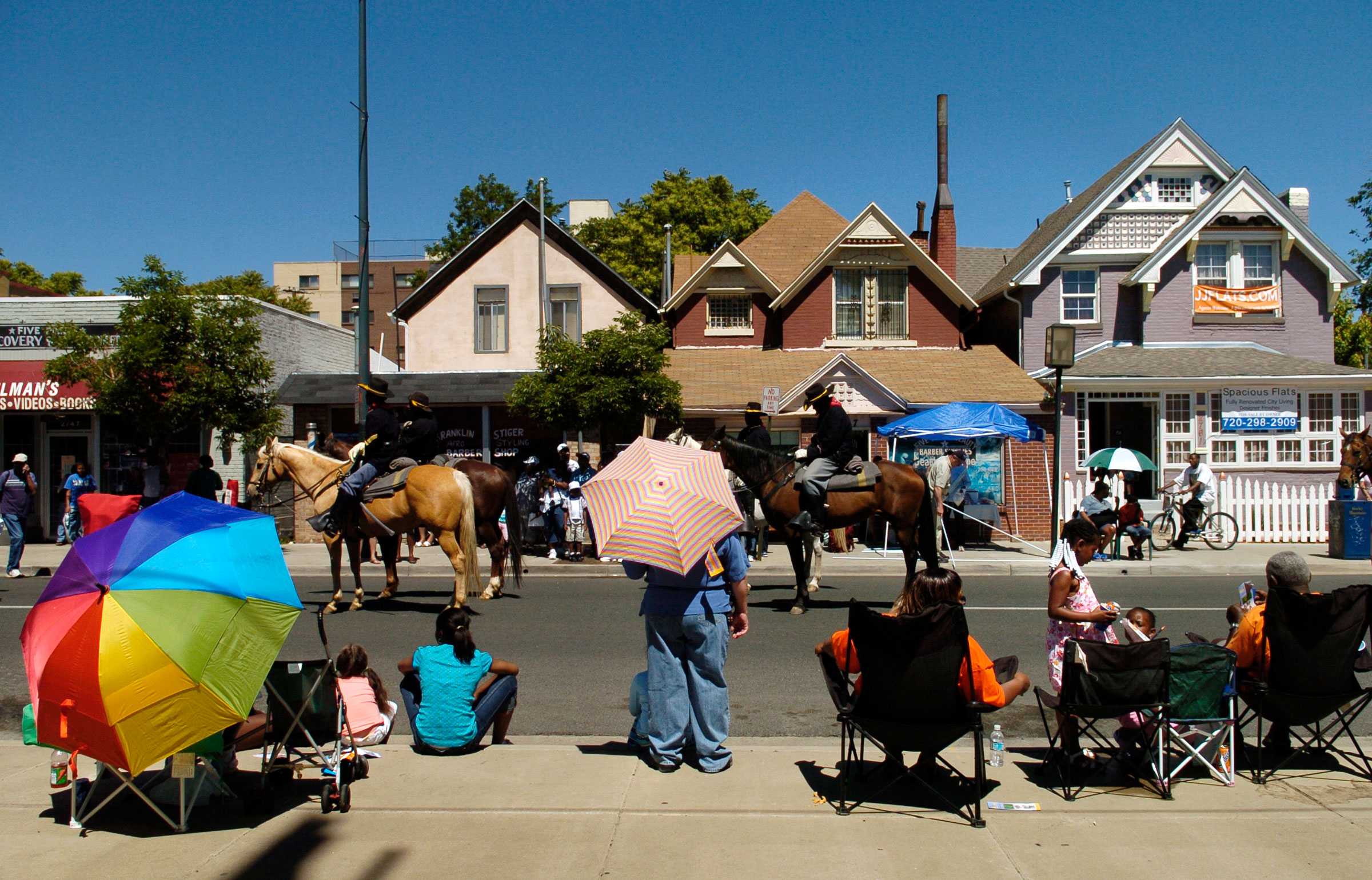 People watch a parade of mounted riders on a sunny day. Colorful umbrellas and old houses line the street.