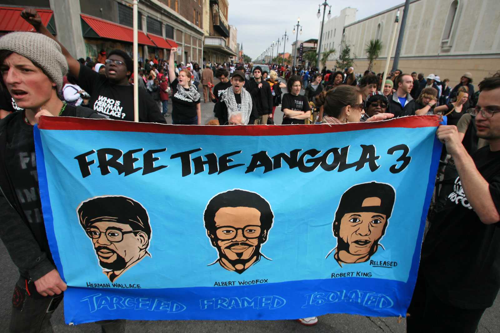 Two people walking in a protesting holding a large blue sign that says "Free the Angola Three".