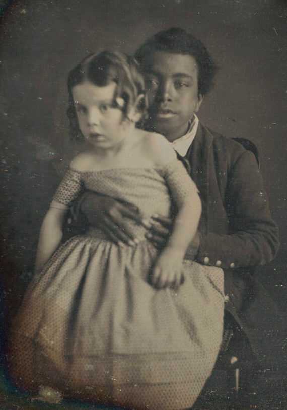 A cased daguerrotype depicting a black boy holding a white male baby on his lap. The baby wears a dotted gown and has hair fashioned into ringlets. The boy wears a dark colored jacket with three bright buttons at the cuff and looks straight ahead at the camera, his hands at the baby's waist. The daguerrotype is in a rectangular gold metal frame with a curved top, and housed in a hinged brown case with red velvet interior and two hook and eye clasps on one edge.