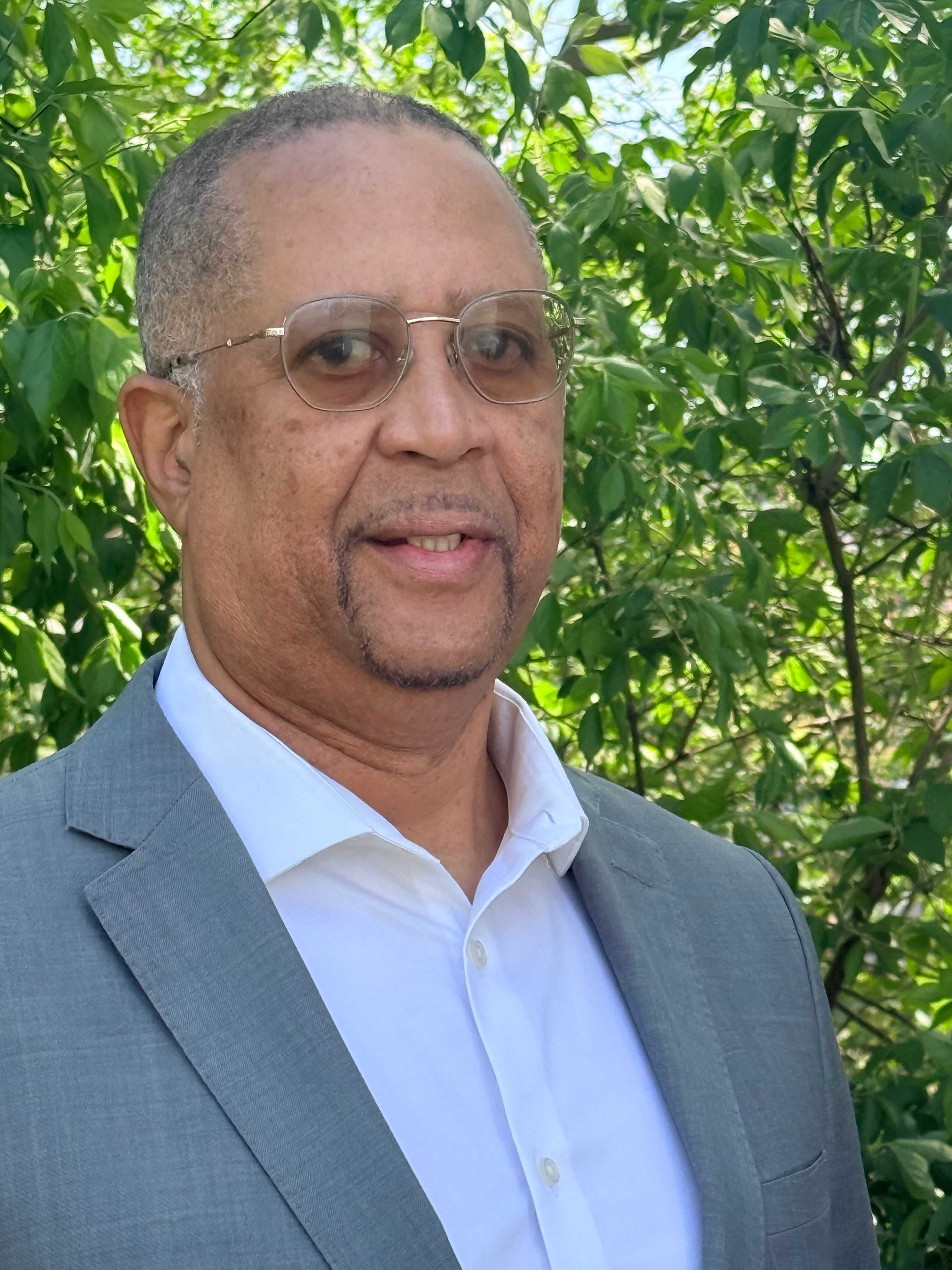 Adult man wearing glasses, a white shirt, and a gray suit jacket, standing outdoors in front of green leafy trees.