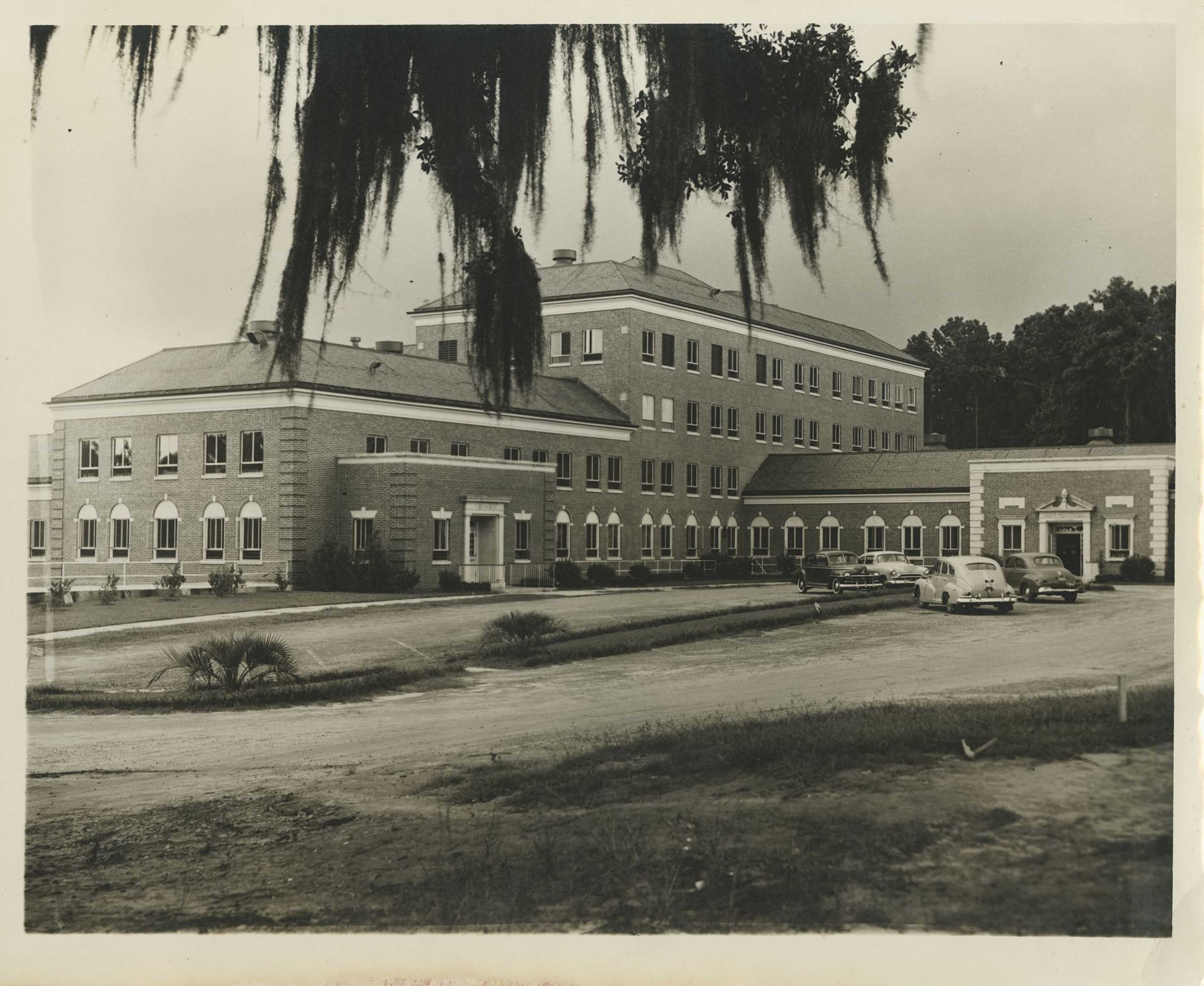 Photograph of a large brick building with cars parked in front, surrounded by trees.