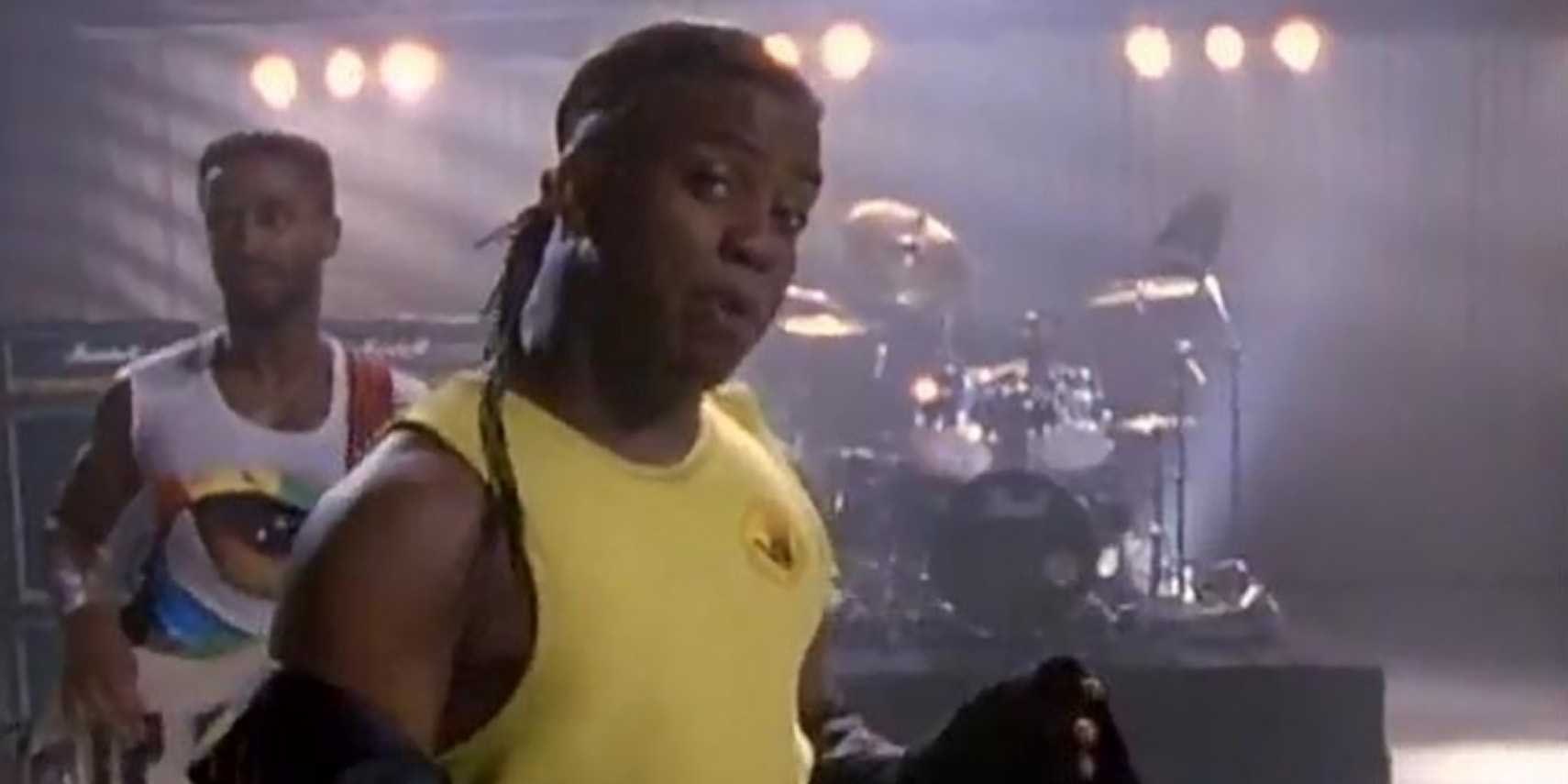 Man in yellow sleeveless tshirt sings into the camera in front of a drum set on a studio.
