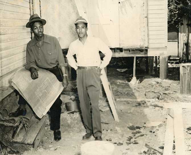 Two individuals at a construction site—one leaning against a building holding a large material, possibly a screen or blueprint, and the other standing with hands on hips. Both wear hats and casual clothing.