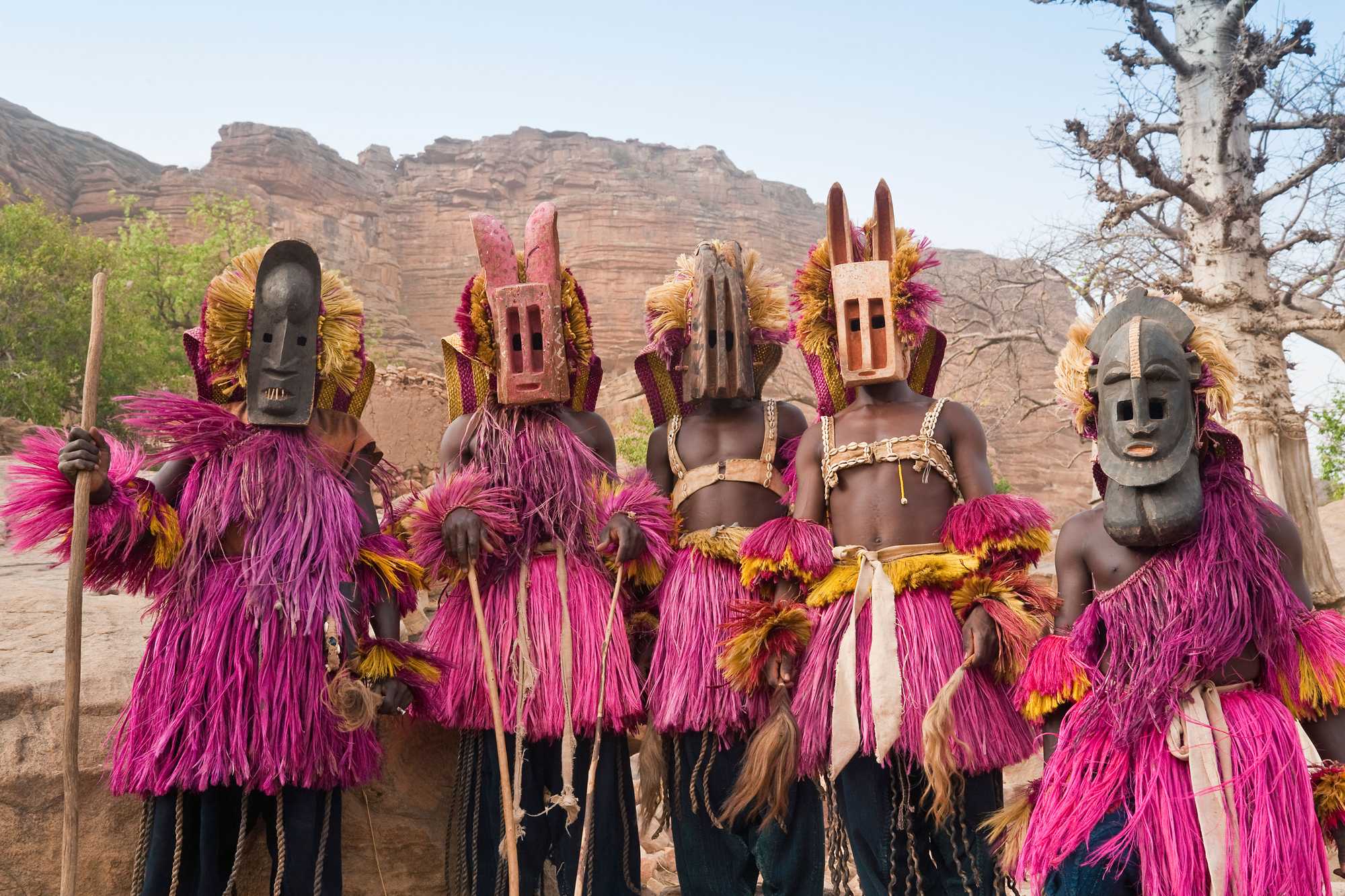 5 dancers stand next two each other. Each has a ceremonial mask and outfit.