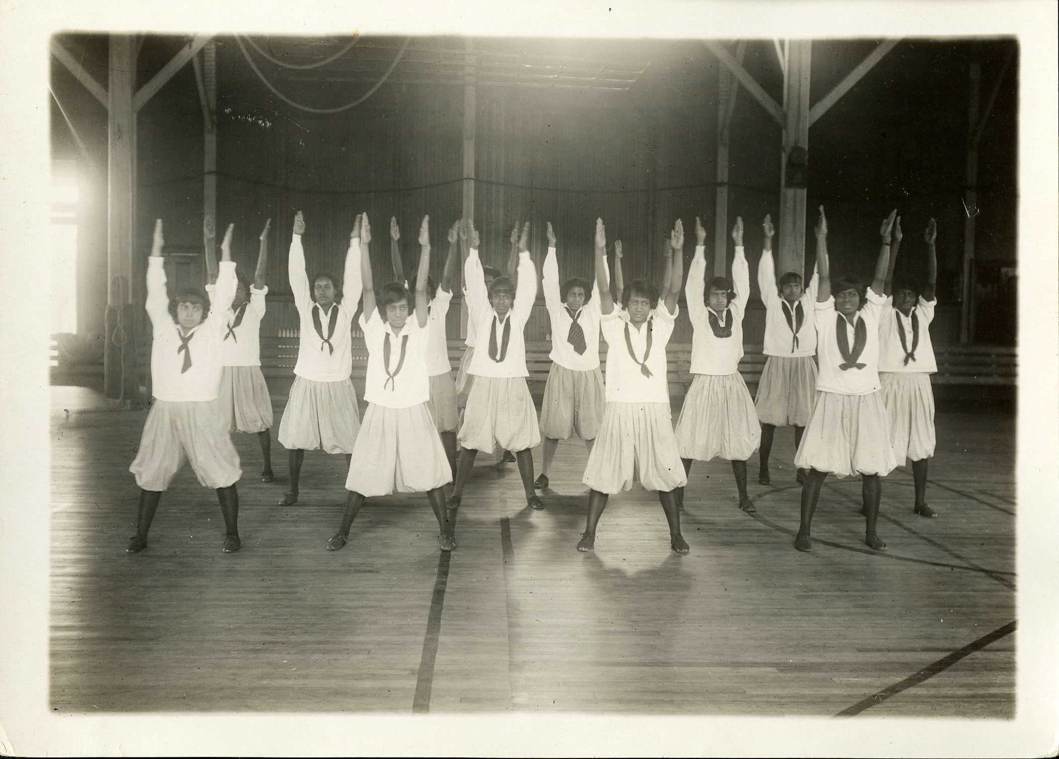 Black-and-white photo of a group of girls in uniforms performing synchronized exercises in a gymnasium.
