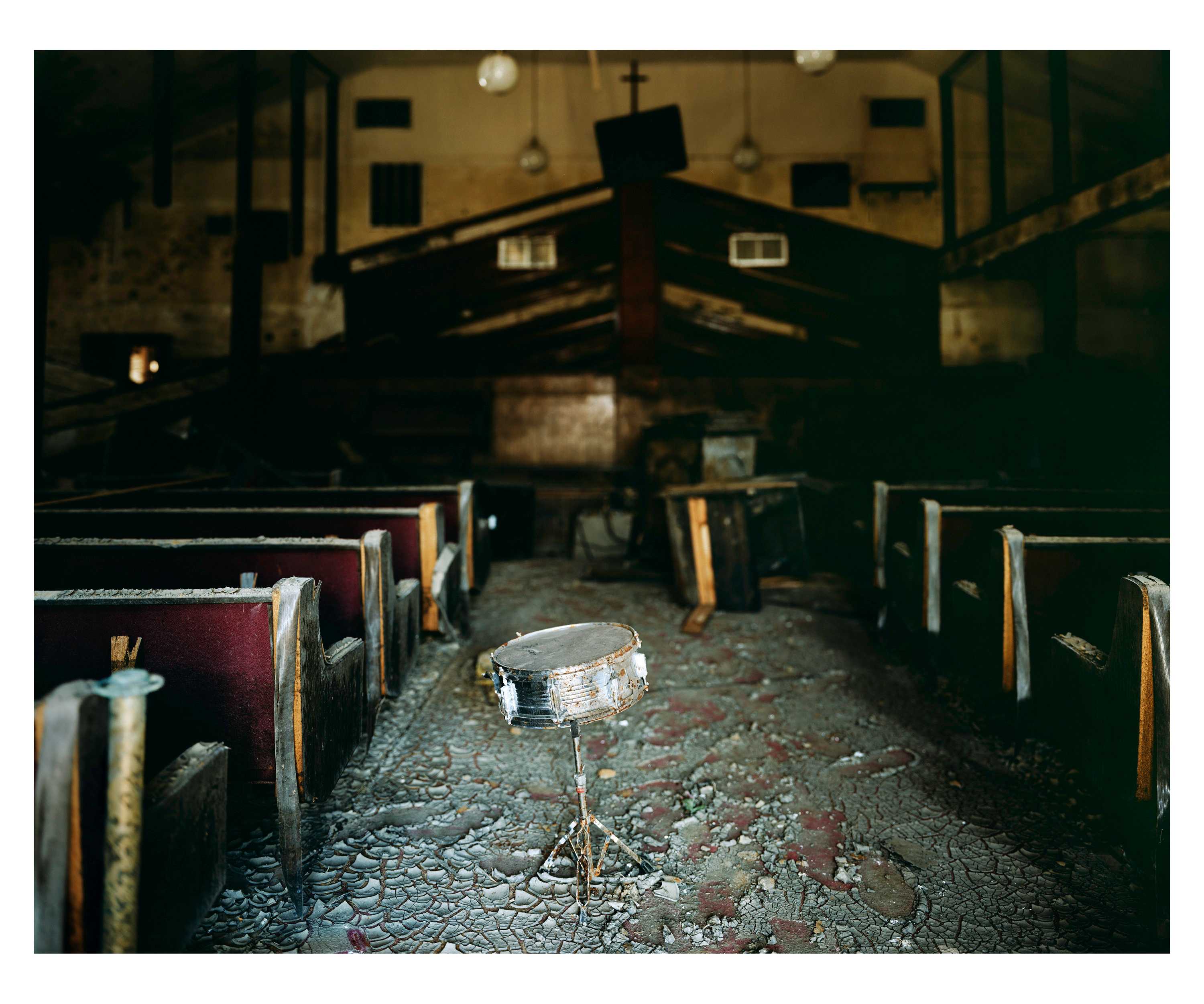 Color photograph of the interior of a church after being flooded by Hurricane Katrina. The image looks down the mud-caked central aisle of the church towards the altar. Sitting in the middle of the aisle is a steel snare drum. To the left and right are pews, and further down the aisle are pieces of debris in front of the dais.