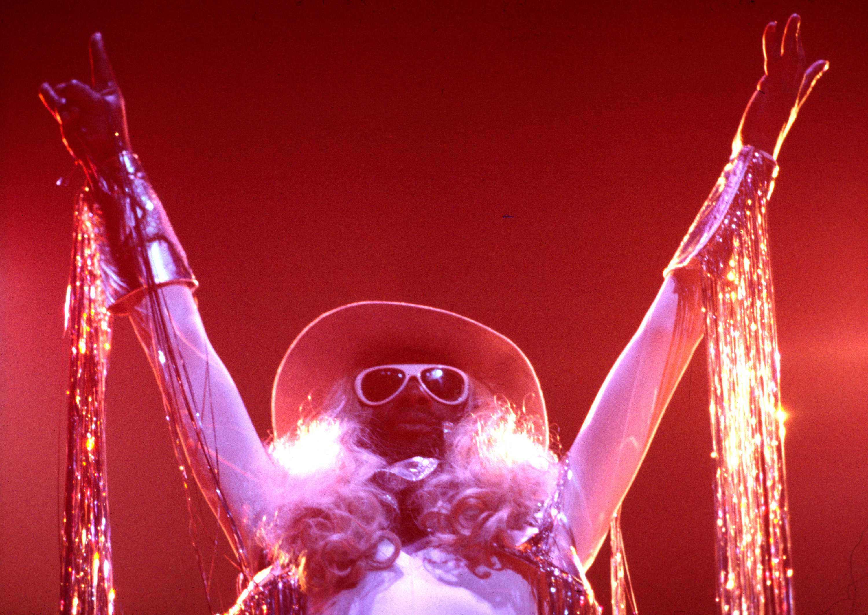 George Clinton striking a powerful pose rocker pose. He is wearing a oversized hat and glasses.