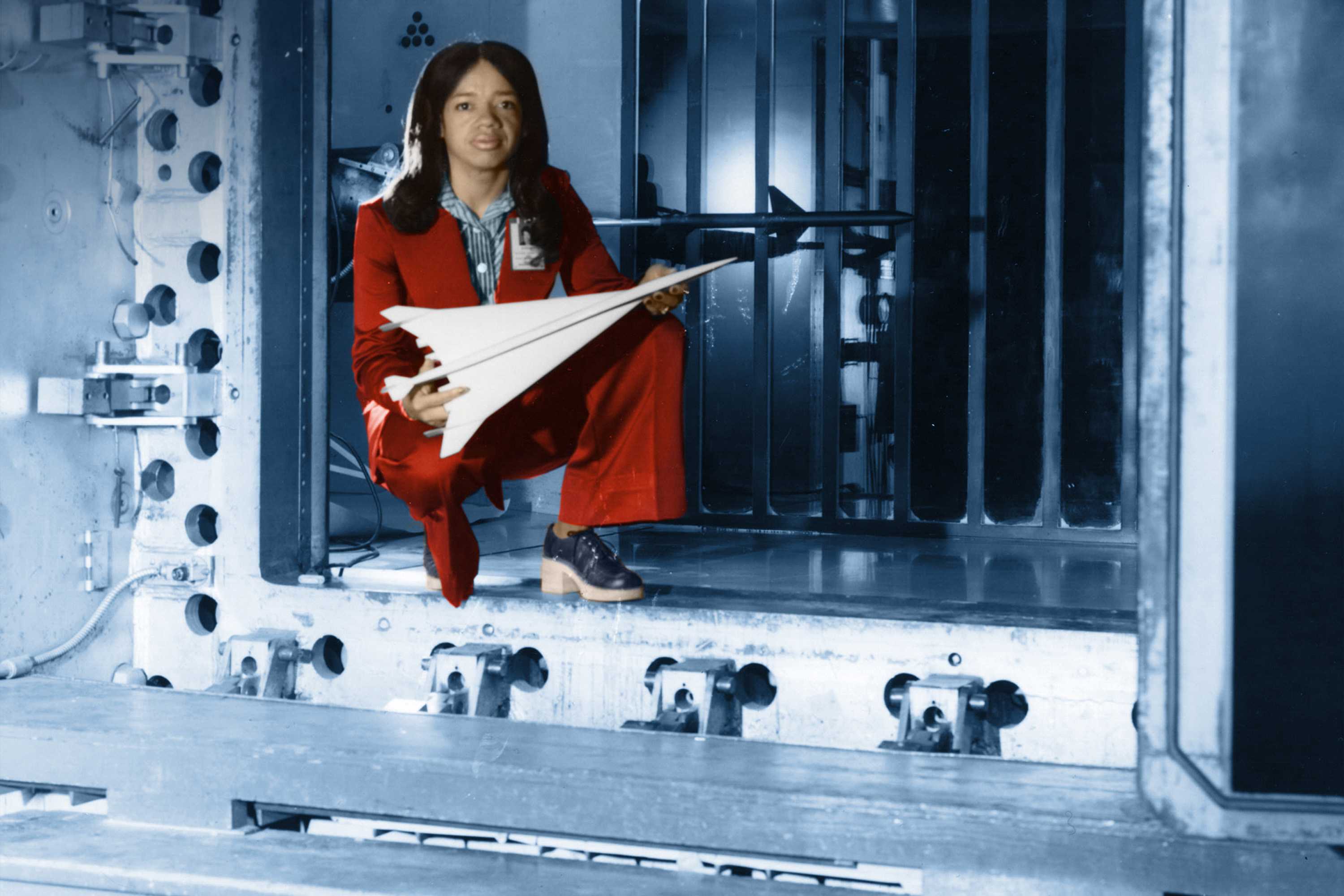 Christine Darden is crouching instead of metal structure as she poses for a photo. She is wearing a red suit.