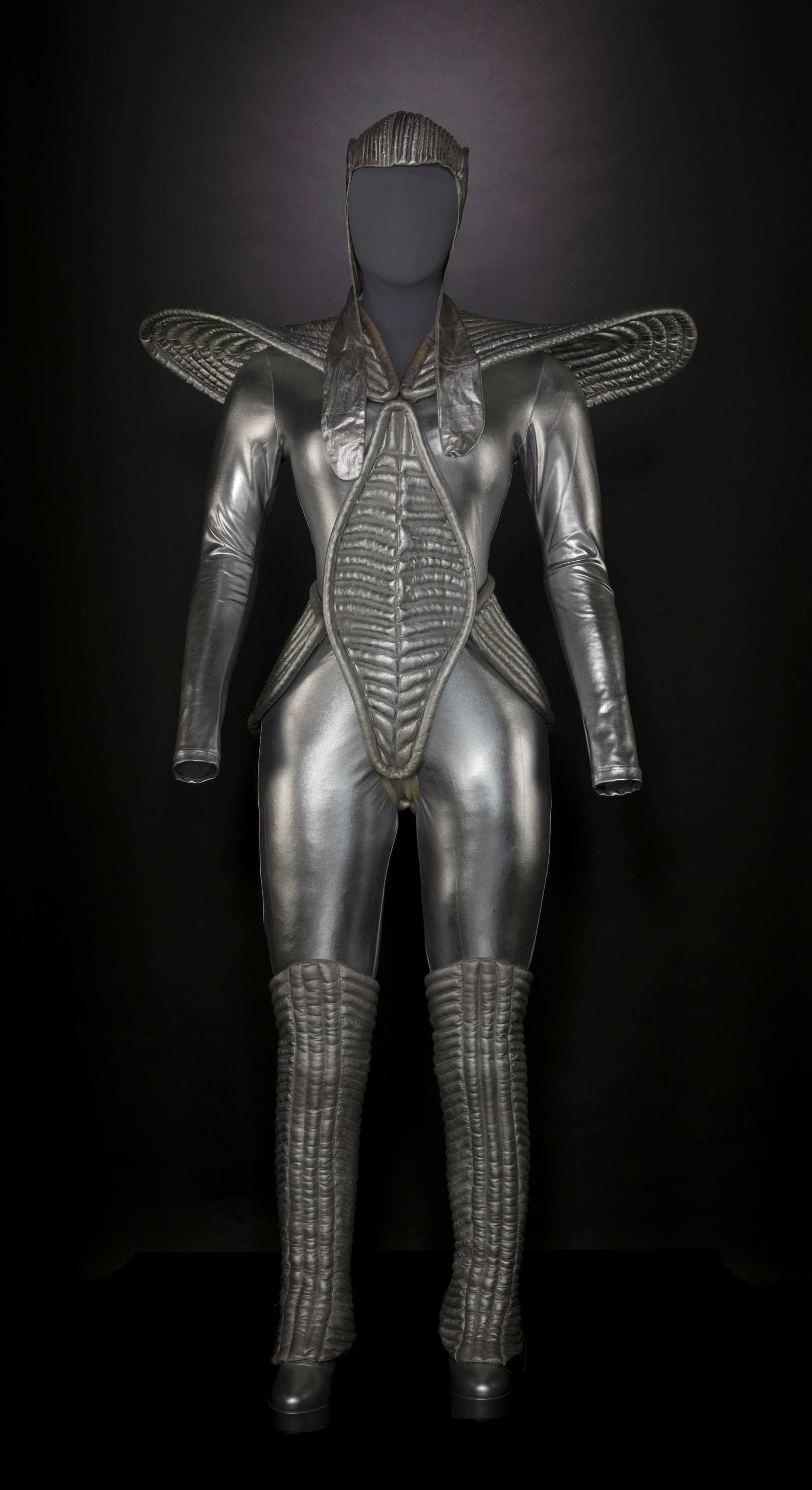 A silver spacesuit like costume covers her head and has oversized shoulder pads.