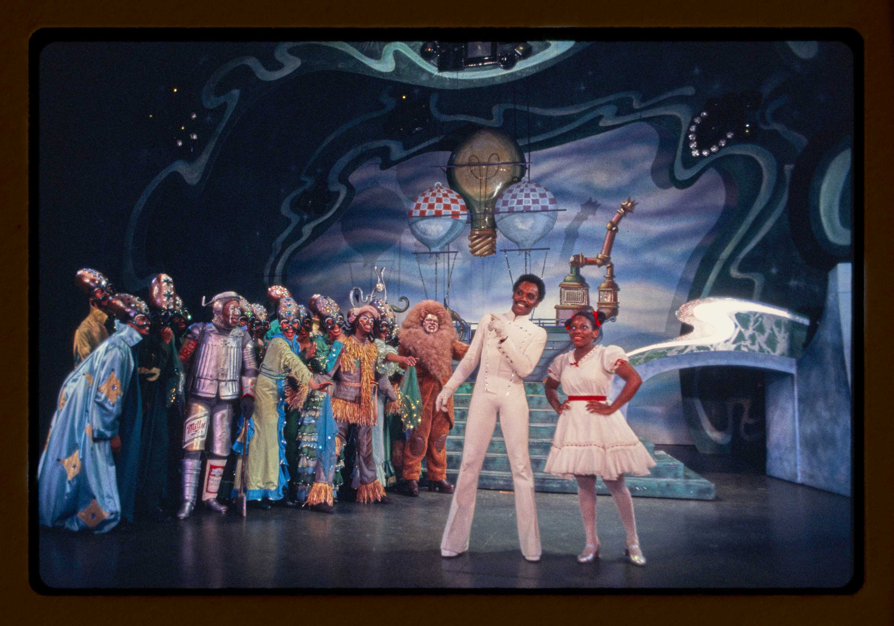 Two characters standing on the right side of the stage in front of other cast members in the Land Oz.