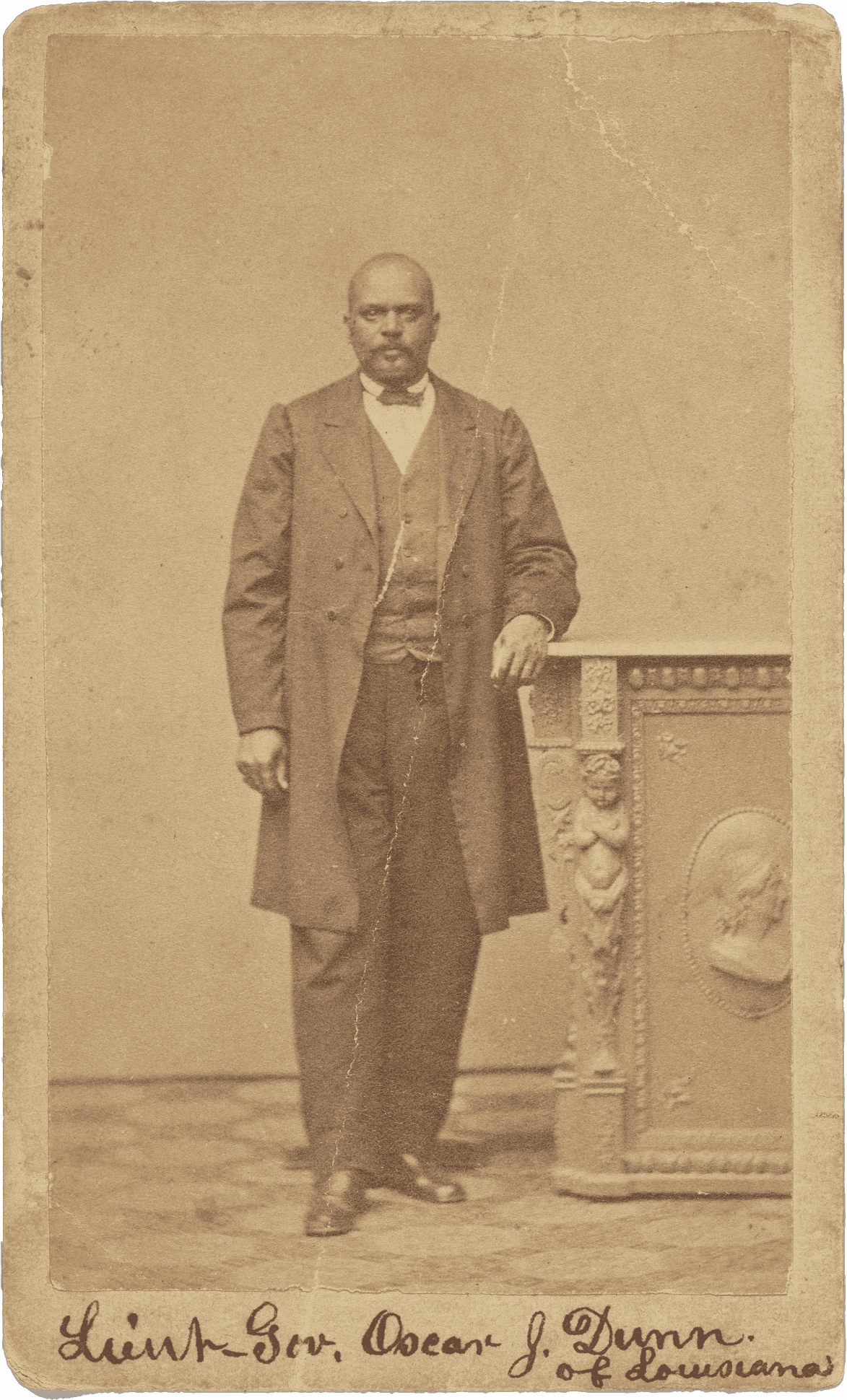 An albumen print carte-de-visite portrait of Lt. Governor Oscar J. Dunn. He is photographed standing with his right foot slightly forward than his left and has his left arm resting on a pedestal. He is wearing a dark colored suit, vest and a bowtie. A watch chain is visible on the right side of his vest. He is looking directly at the camera. The photograph is inscribed on at the top and bottom of the front. At the bottom, handwritten in black ink, is: [Lieut Gov. Oscar J. Dunn / of Louisiana].