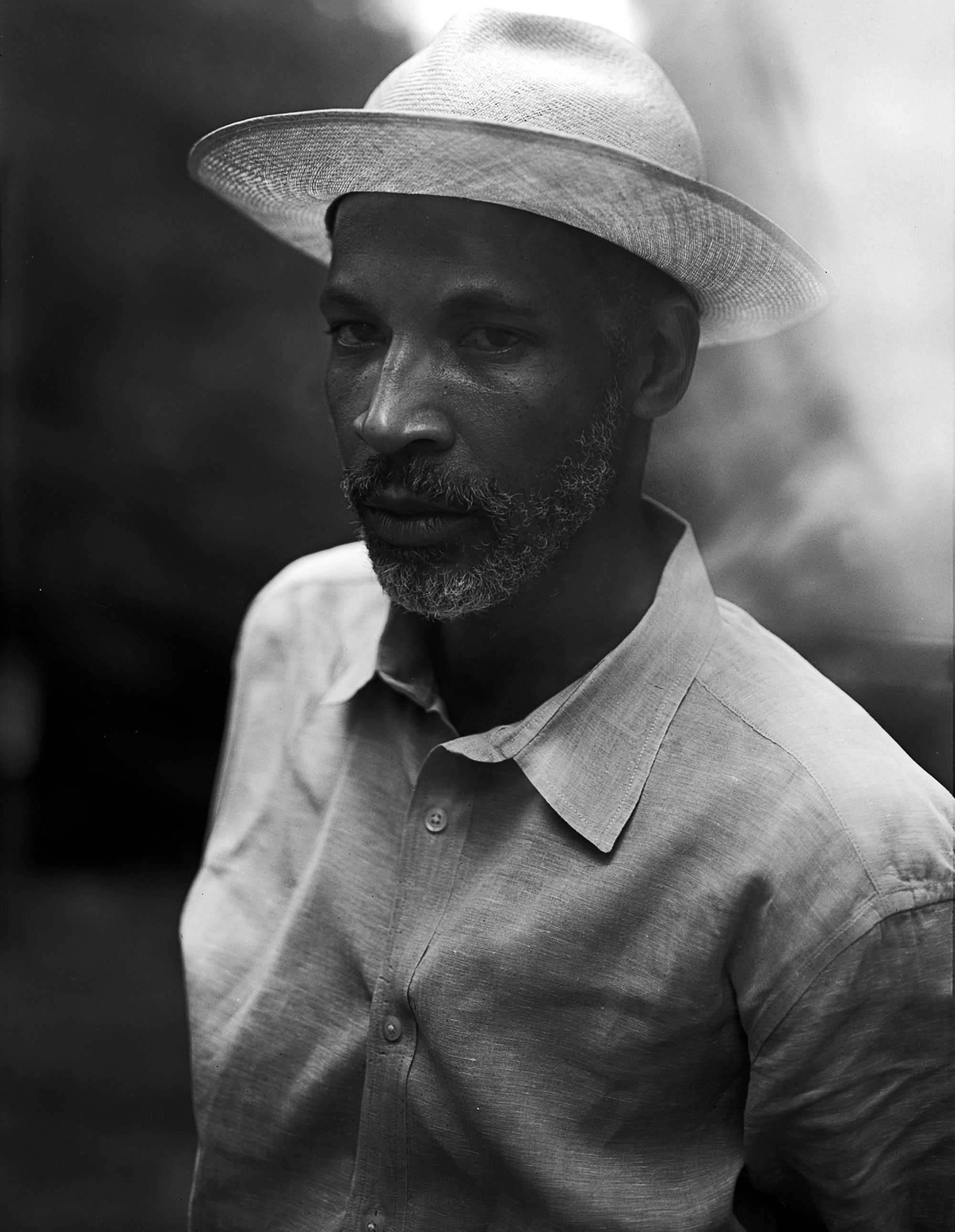 Black-and-white photo of a man wearing a hat and a button-down shirt, standing outdoors and looking toward the camera.