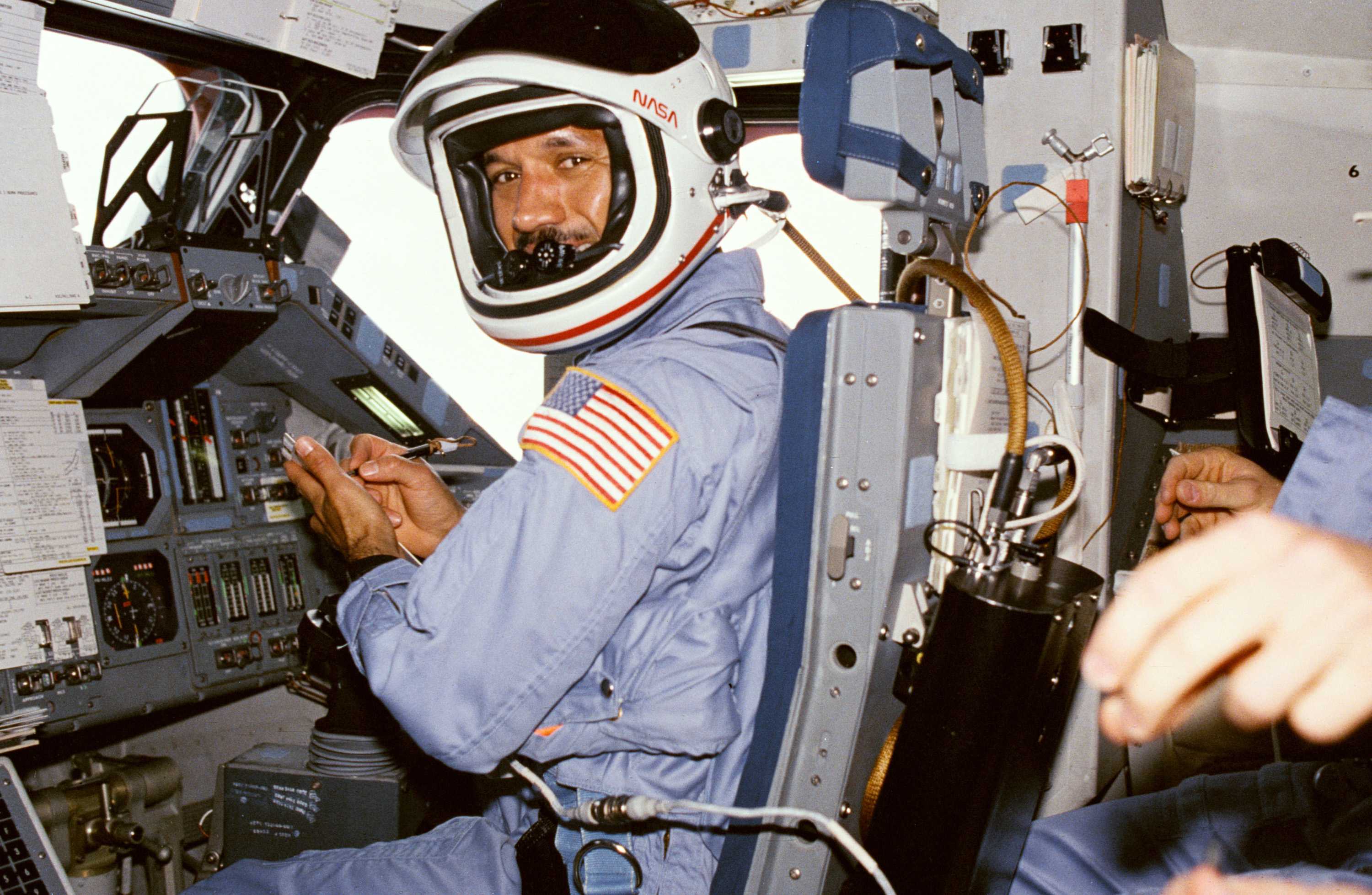 Major General Charles sitting in the pilot seat, dressed in a light blue space suit, with a pen in his hand.