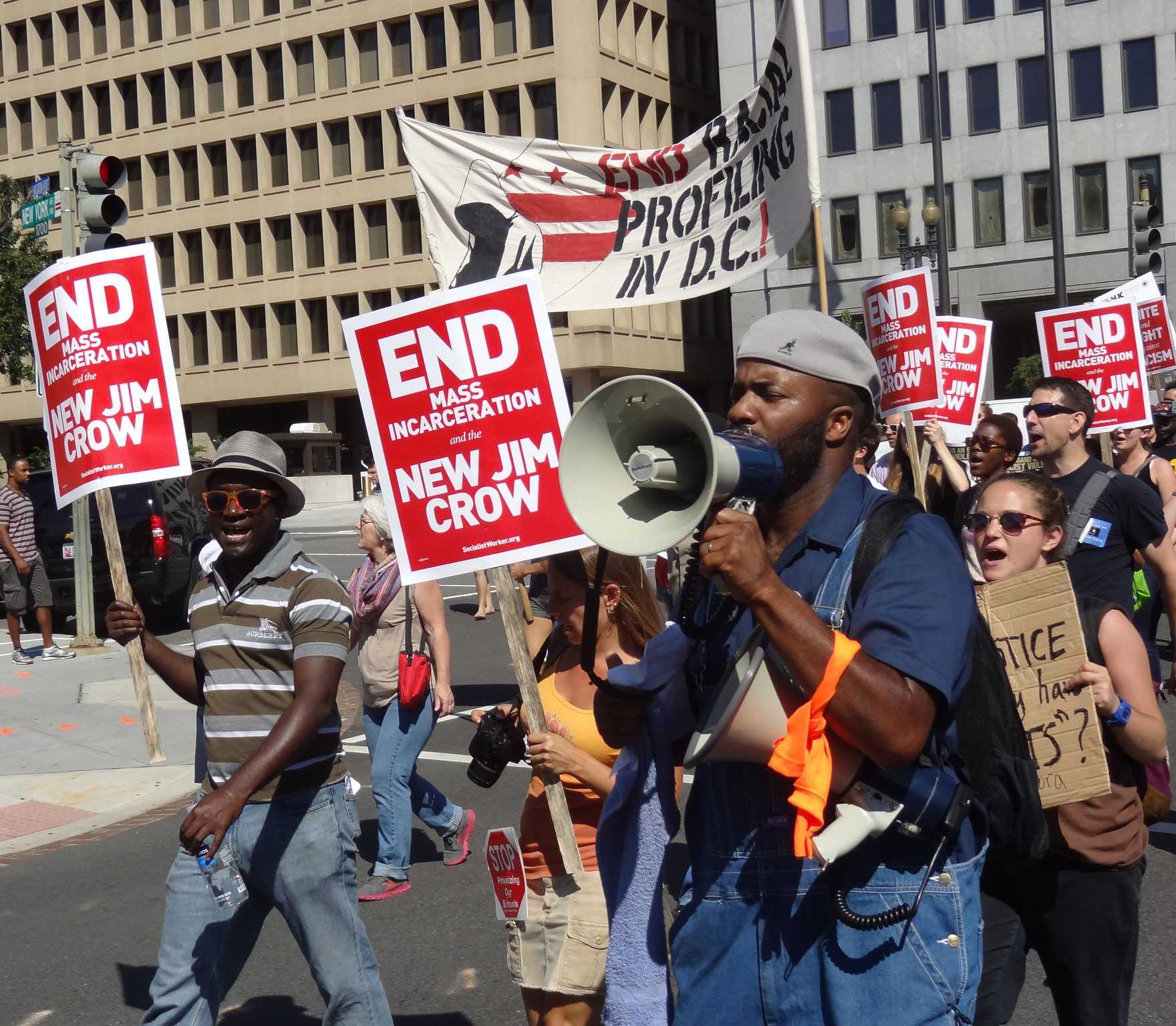 A group of people protesting, walking through a street, with bright red signs that say 'End Mass Incarceration'