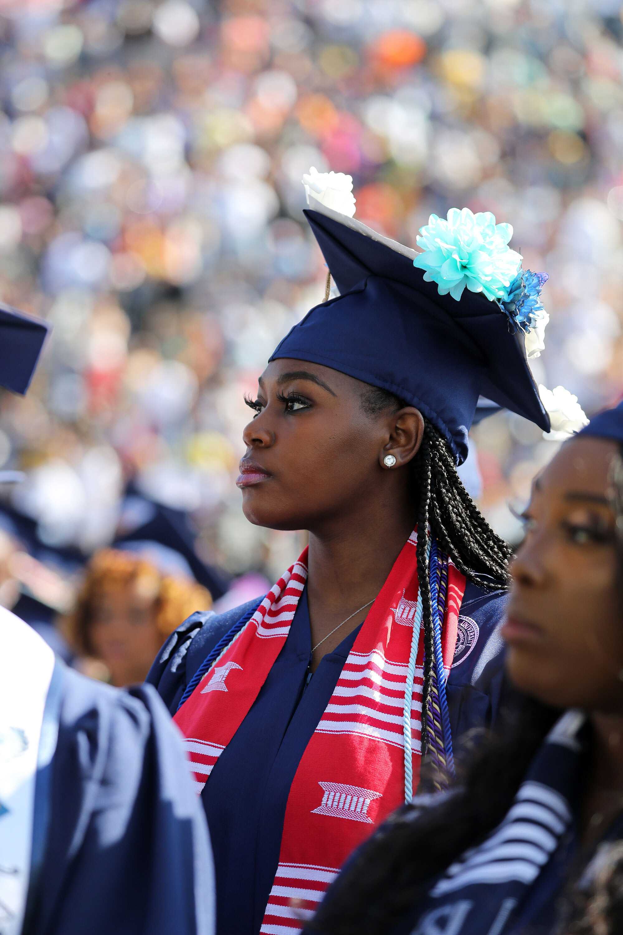 A graduate wearing a cap and gown with a decorative cap stands among fellow graduates at a ceremony.