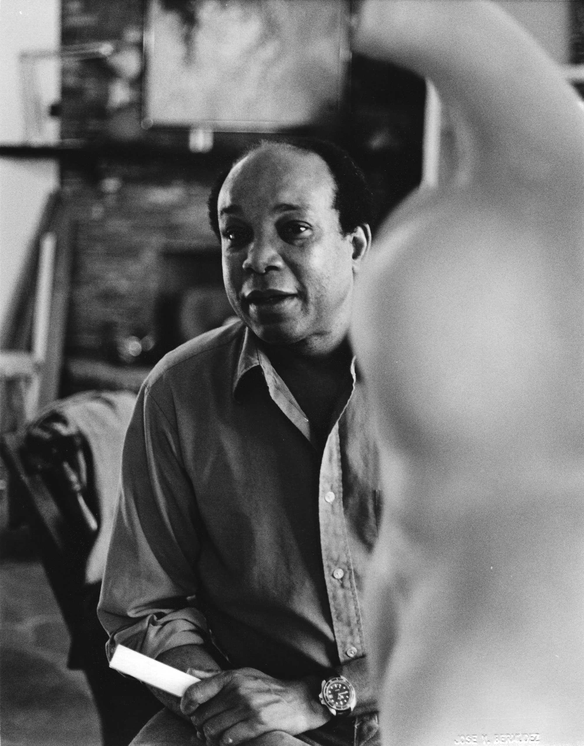 Black-and-white photo of a man sitting with a sculpture in the foreground.