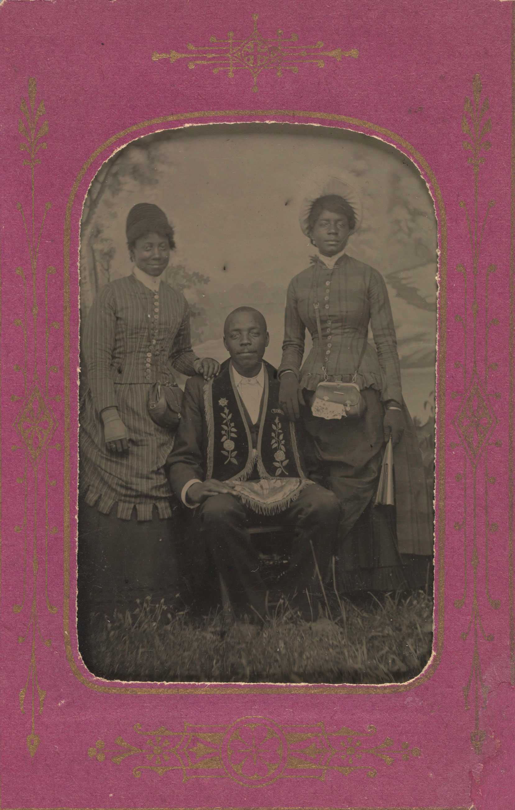 A tintype of two women standing next to a sitting man in a pink decorative frame.