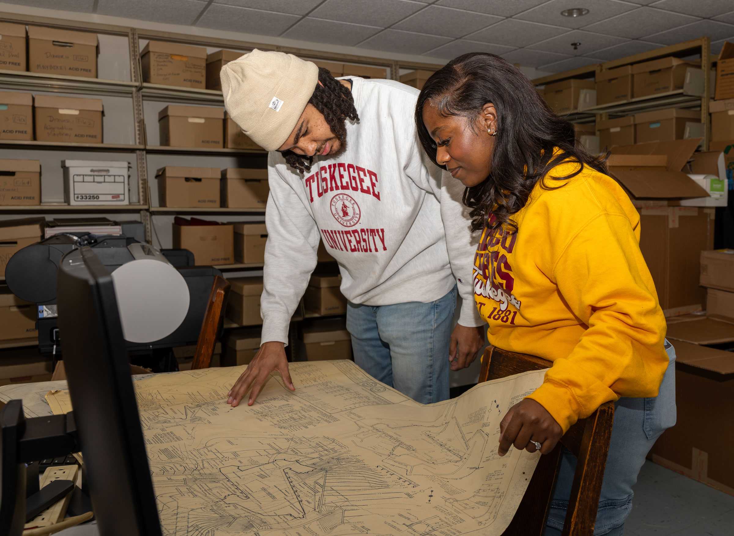 Color photograph of two people reviewing a large architectural plan in a storage room with boxes and equipment.