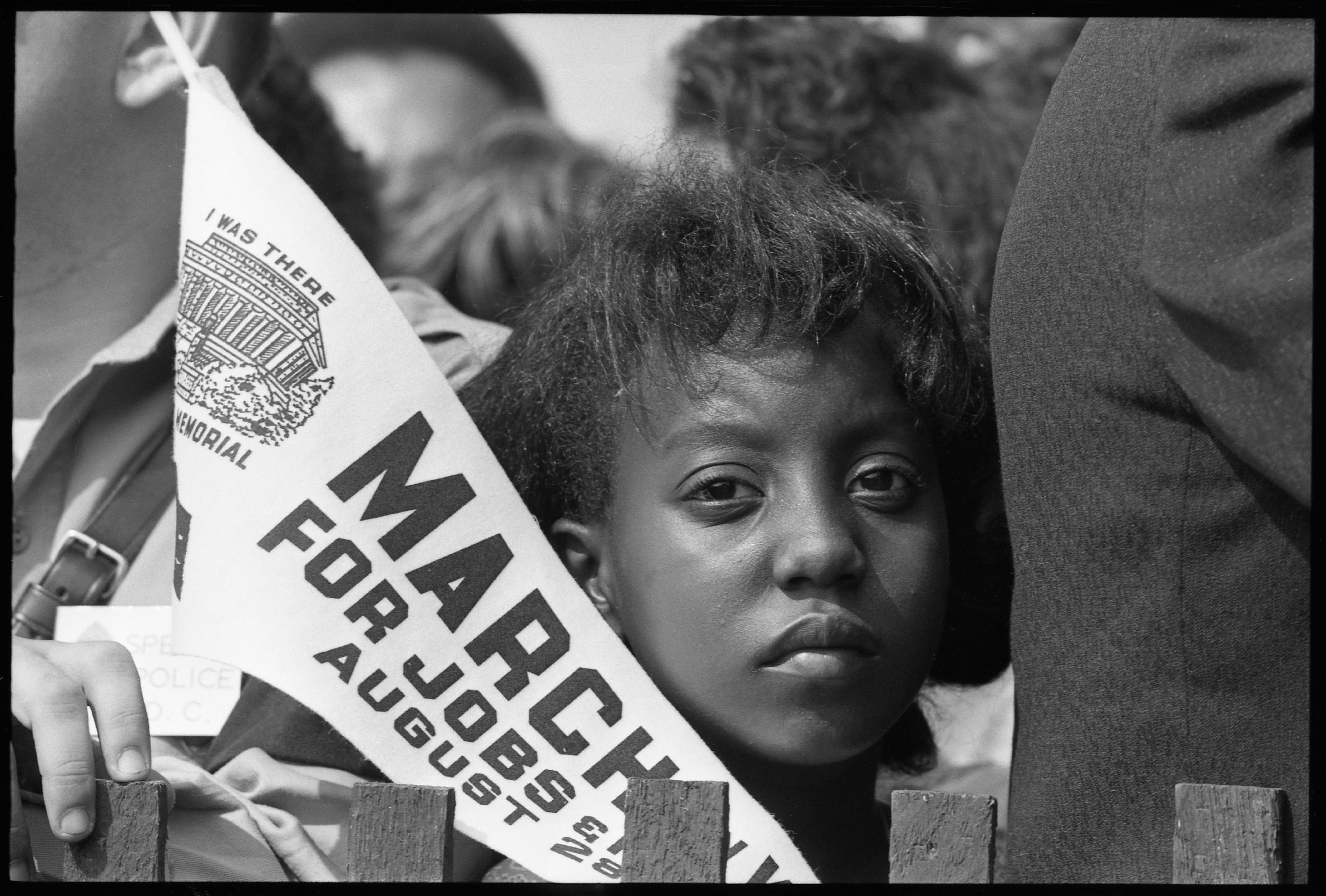 Black-and-white photo of a child in a crowd holding a ‘March on Washington for Jobs and Freedom’ pennant, with people closely gathered behind a fence.