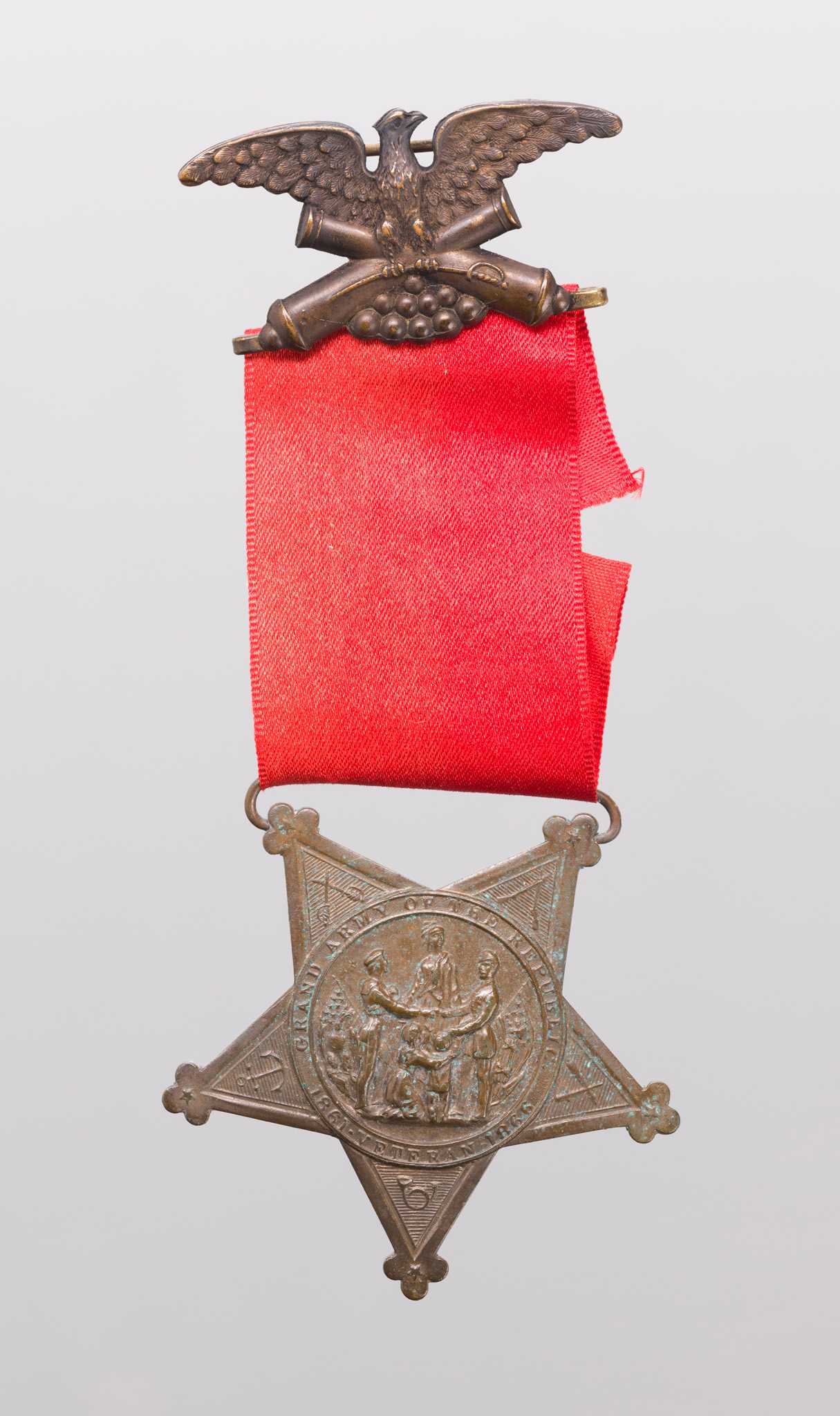 A Grand Army of the Republic medal owned by Marquis Peterson. The medal has a suspension brooch in the shape of an eagle with spread wings above crossed canons. It is attached to a red ribbon. A metal suspender attaches the medal to the ribbon. The medal is in the shape of a five-pointed star. The front of the medal Has the seal of the Grand Army of the Republic at center. The seal shows a sailor and soldier next to American flags. They are shaking hands in front of a standing woman. A kneeling woman and a standing child with raised arms are between the sailor and soldier. Embossed lettering circles the seal around the edge and reads “GRAND ARMY OF THE REPUBLIC” at top and “1861 VETERAN 1866” at the bottom. Around the seal on each point of the star additional emblems are embossed. They are crossed sabers, crossed rifles, crossed cannons, a bugle and an anchor. The back of the medal has a shield at center. This is surrounded by 8 fraternal symbols forming a circle. This circle is surrounded by another circle of 16 fraternal symbols. This is encircled by 34 five-pointed stars. The five-star points on the back of the medal also have things embossed on them. The top two connecting to the suspender ae olive branches. The tow on the side are wreaths and the point on the bottom is of a banner.