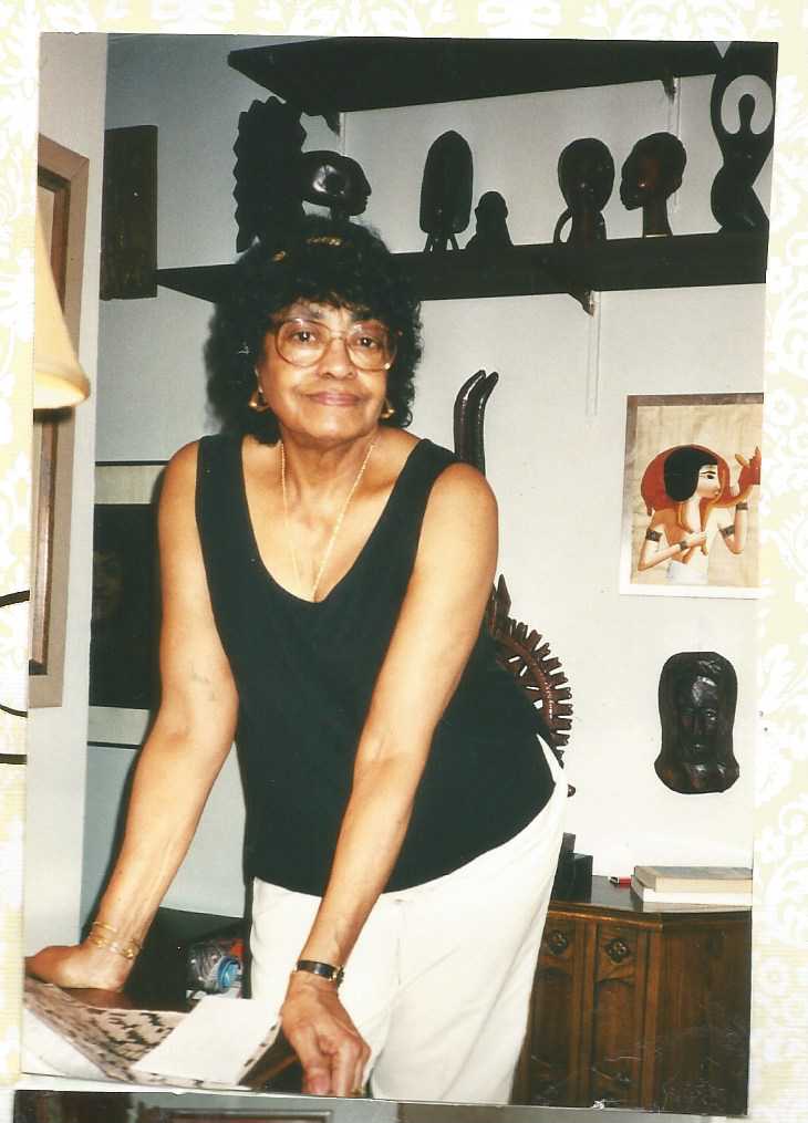 Color photograph of African American woman leaning on her desk.  There are small sculptures on a shelf behind her and other art work on the walls.