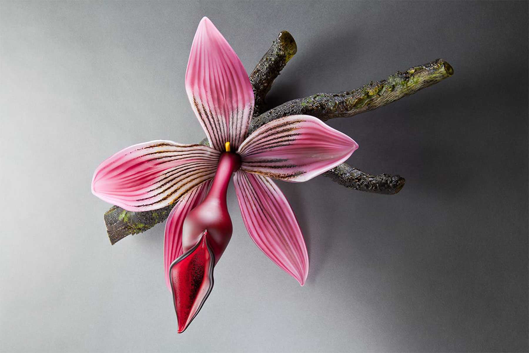 A glass sculpture of a pink orchid with detailed petals, displayed on a textured branch against a gray background.