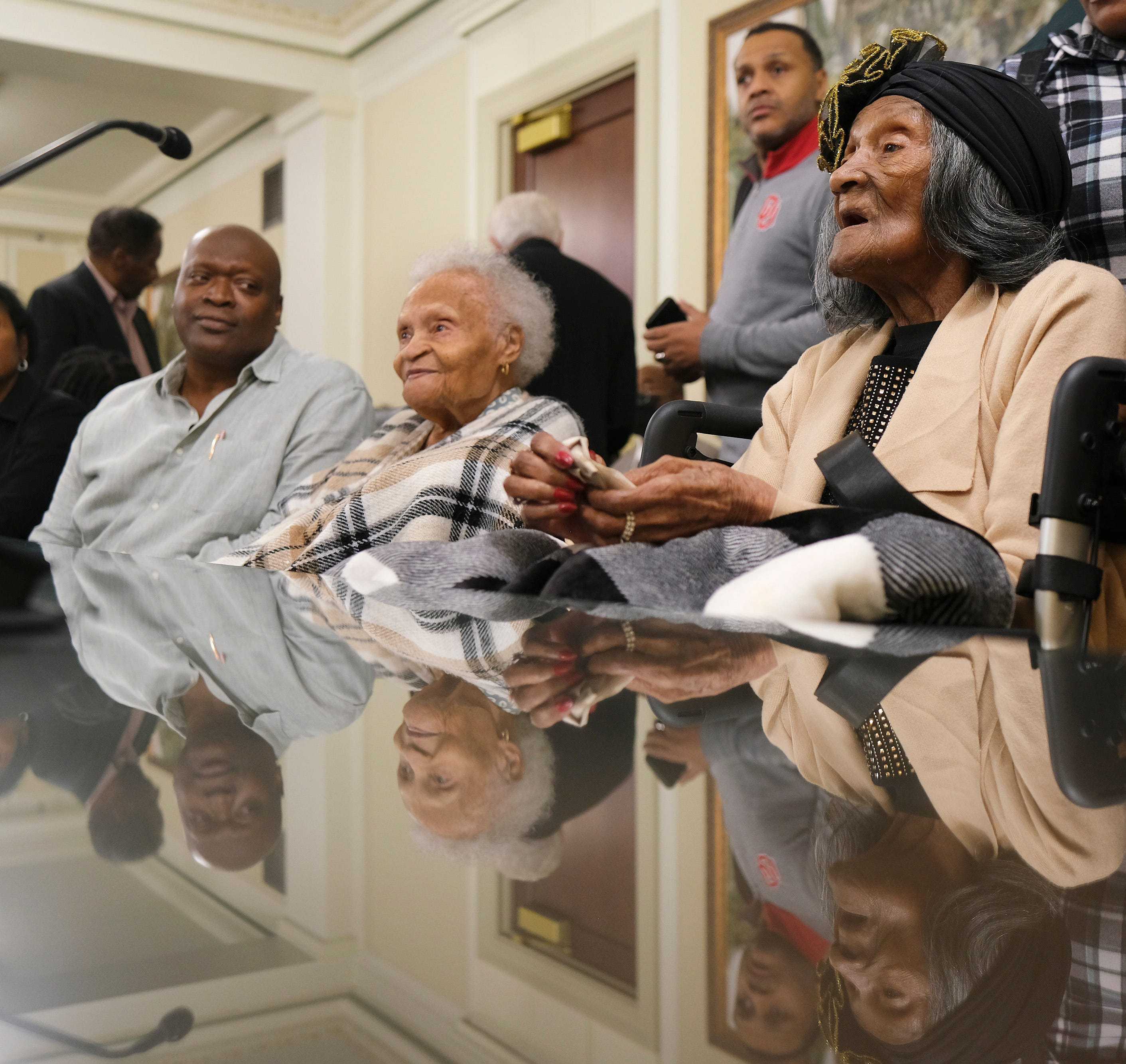 Color photograph of two elderly women and man sitting at table.  There is a microphone in view and people standing behind the three.