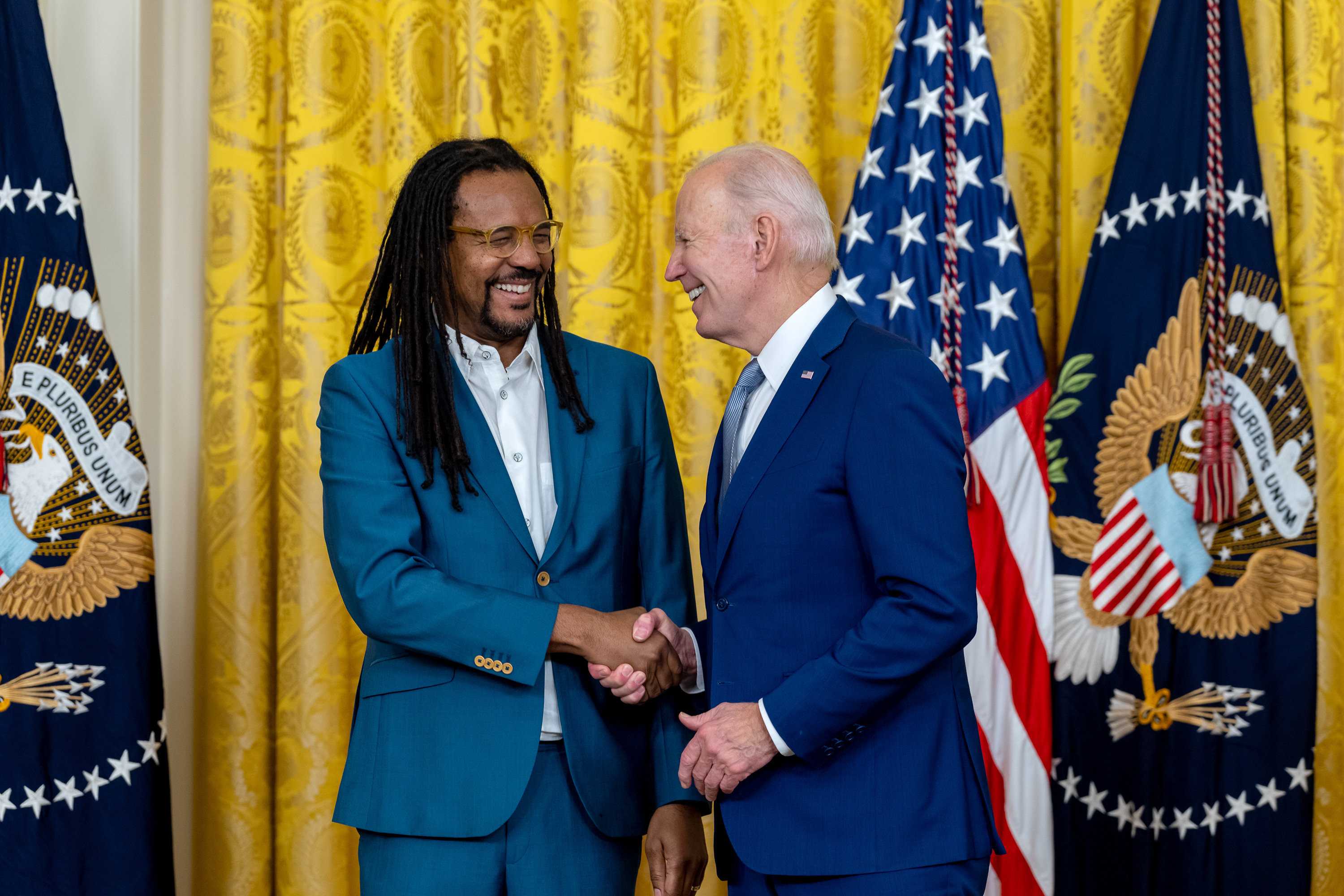 Colson Whitehead and President Joe Biden shake hands in front large gold curtan and american flags.