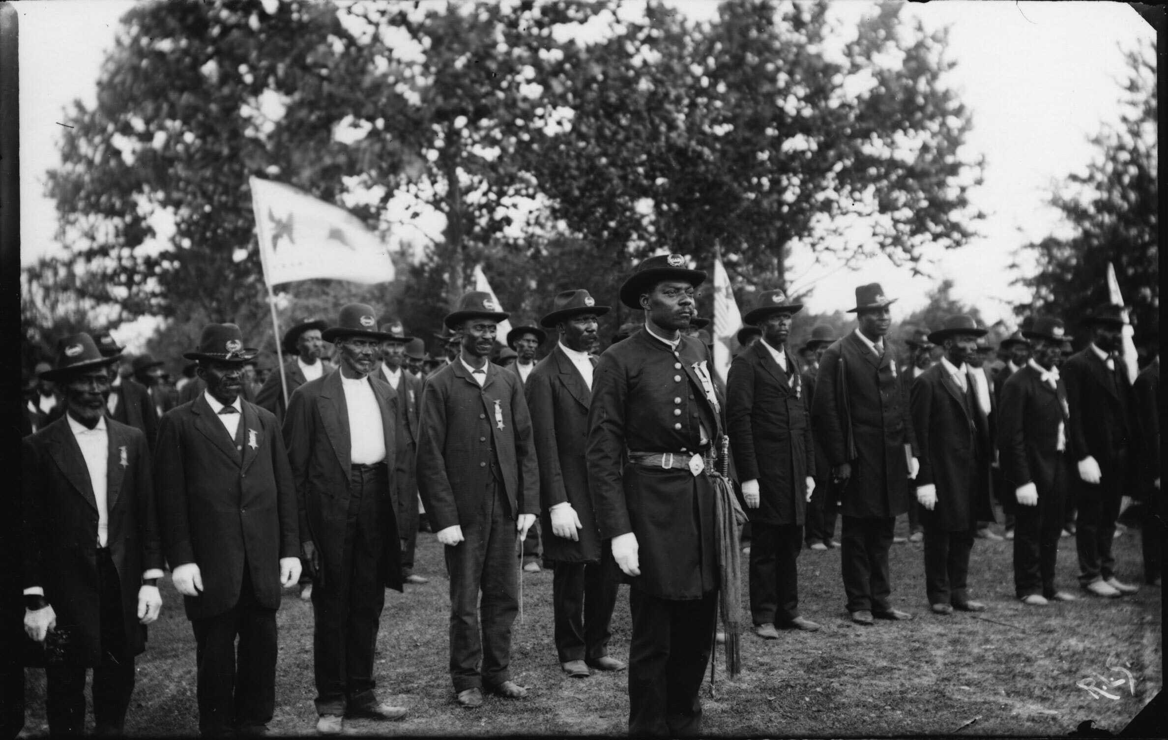 A black and white photo of 2 rows of African American soldiers lined up next to each other. Some hold guns and flags.